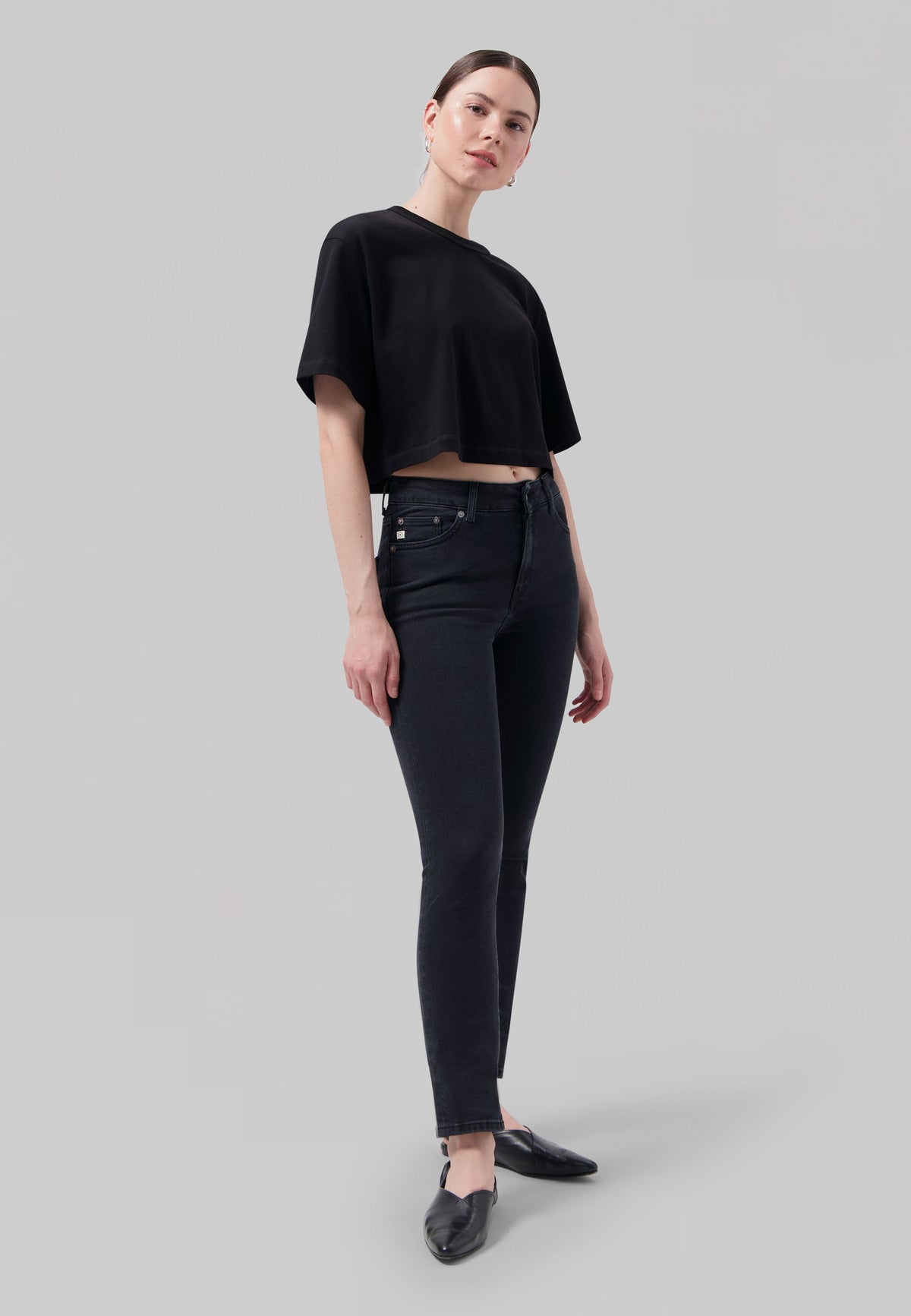 Woman in a black crop top and slim-fit dark jeans standing confidently against a light gray backdrop.
