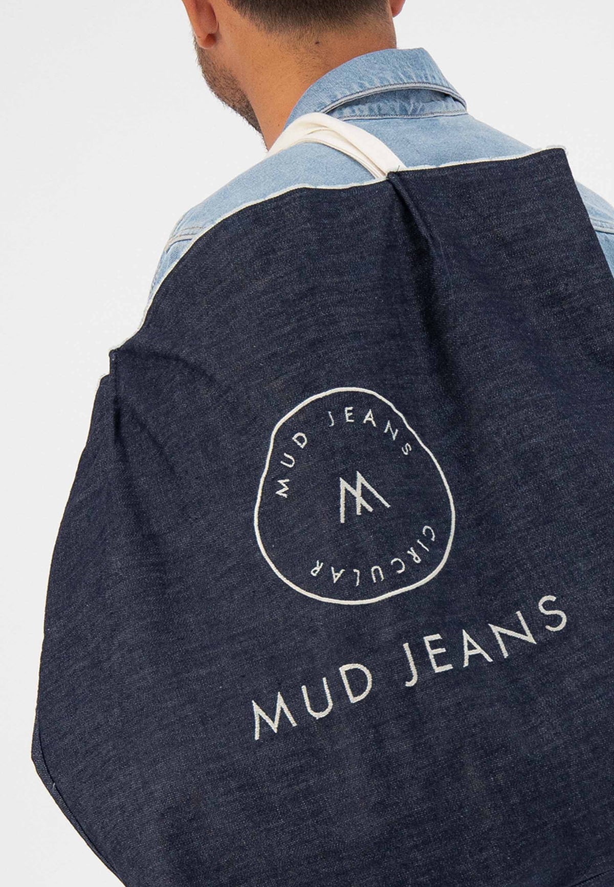 Man in blue denim jacket carrying a dark blue, sustainable MUD Jeans tote bag, featuring circular logo design.
