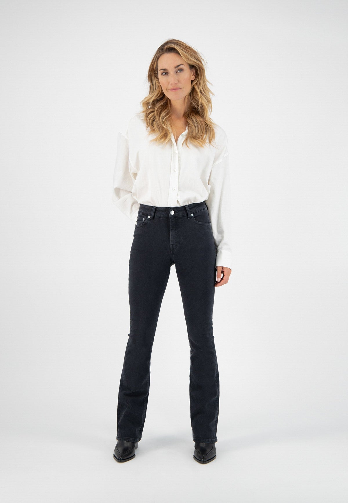 A woman wearing a white button-up blouse and black flared jeans paired with black pointed-toe boots against a plain background.