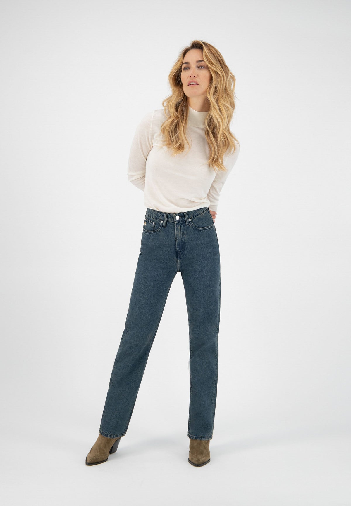 Woman wearing high-waisted blue jeans and an ivory long-sleeve top, posing with hands behind her back.