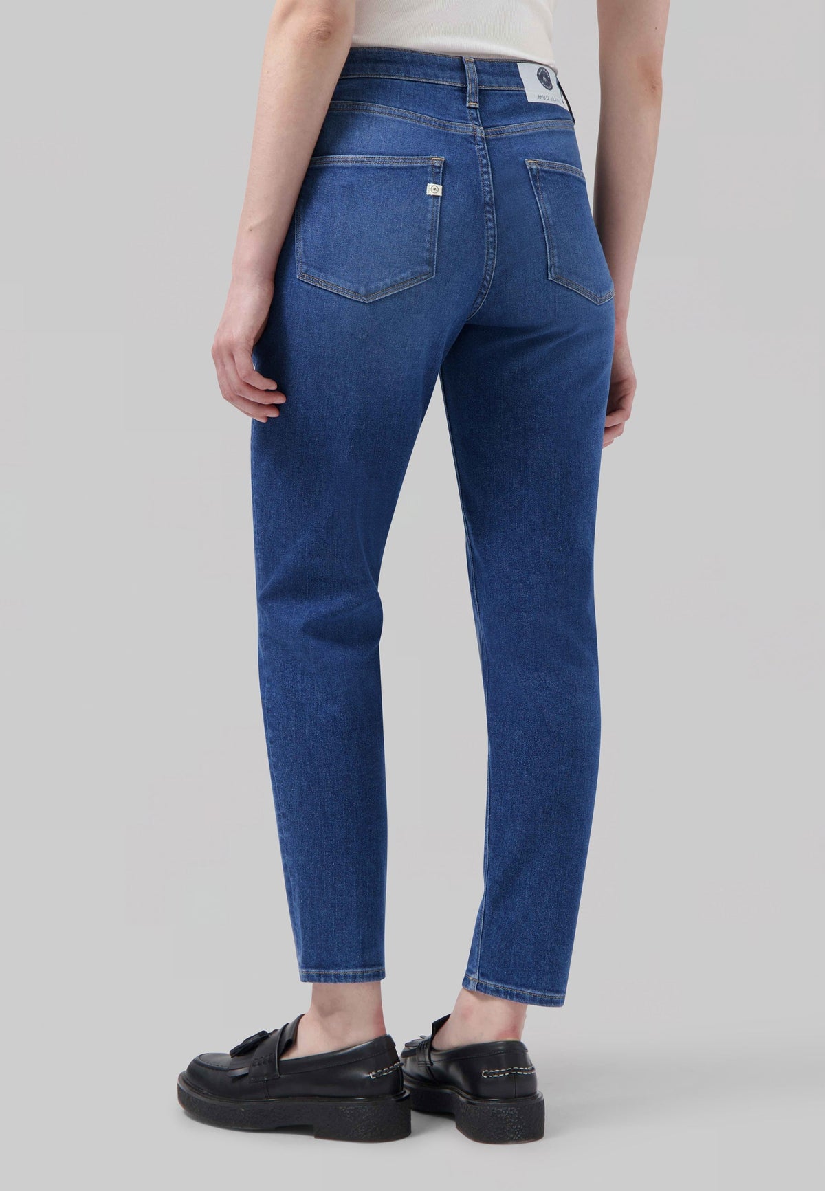 Back view of eco-friendly MUD Jeans, slim-fit, medium blue denim, shown with black loafers and a white sleeveless top