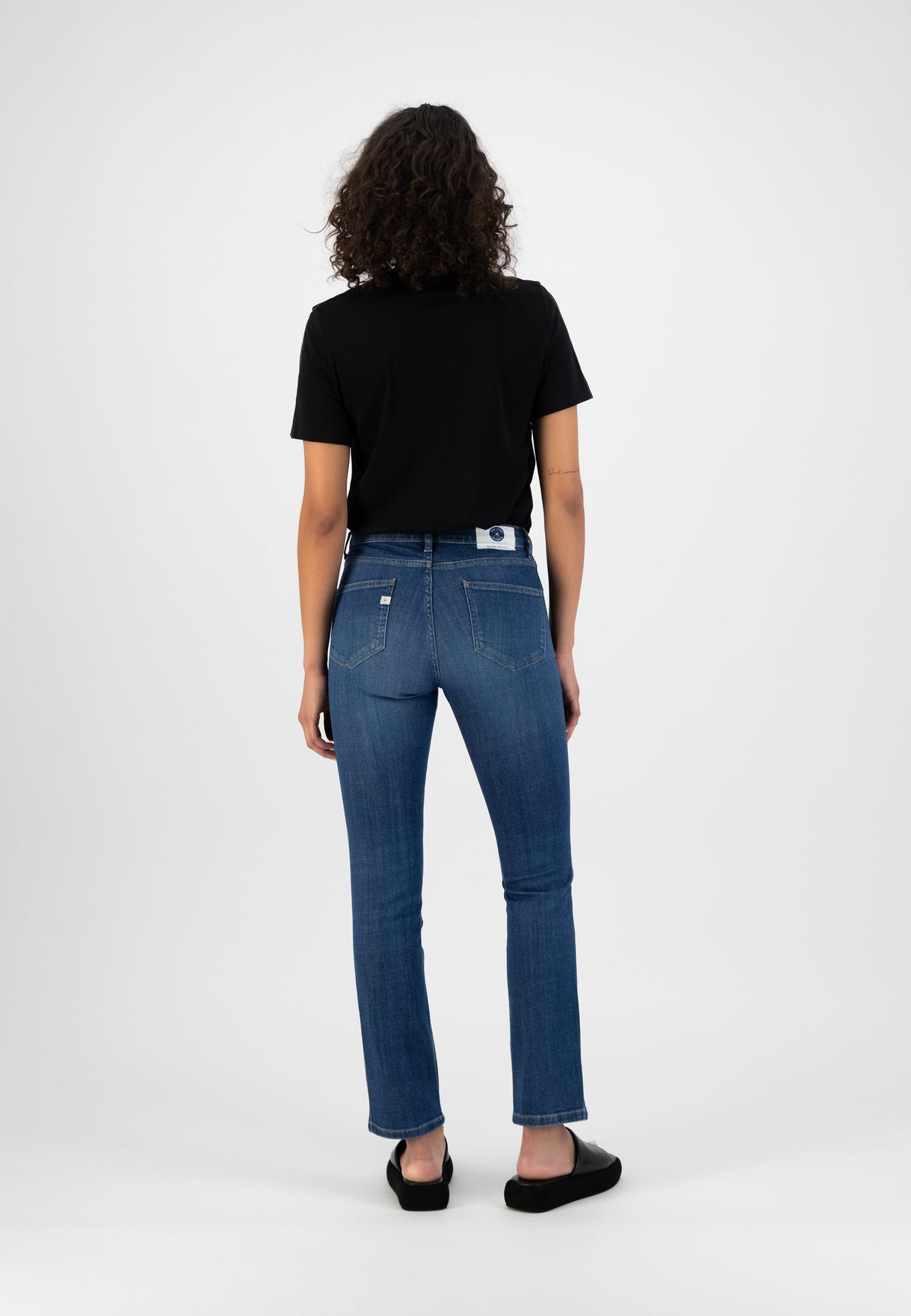 Curly-haired woman in black T-shirt and slim-fit blue jeans, showcasing back view of casual outfit with black slip-ons.
