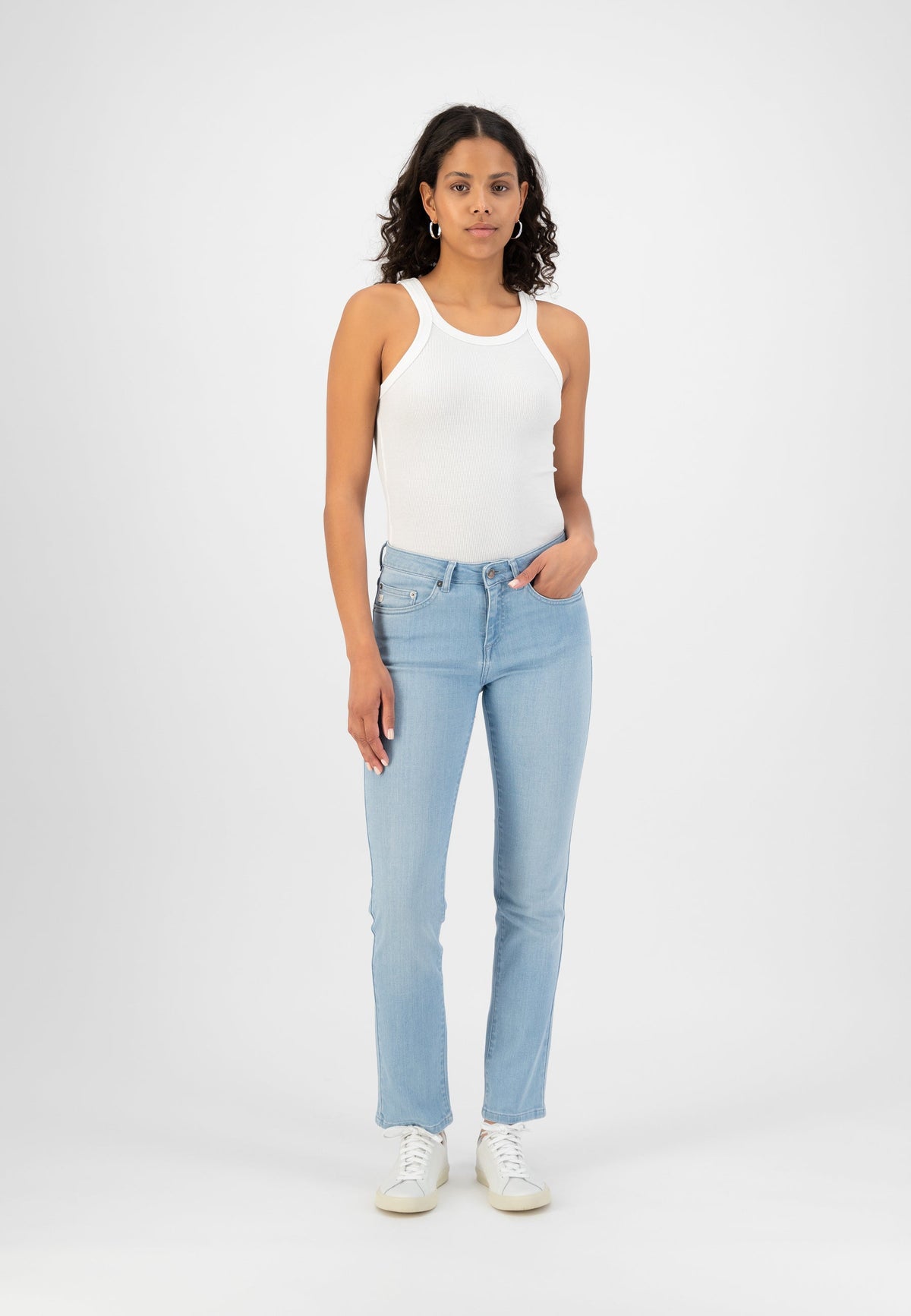Woman in light blue slim-fit jeans and a white ribbed tank top, paired with white sneakers, standing against a plain background.