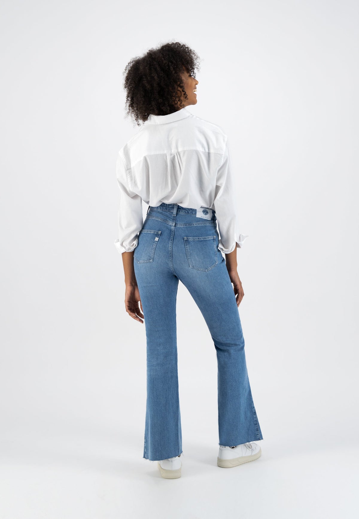 Model wearing a white button-up shirt and high-waisted blue flare jeans, with sneakers, viewed from the back.