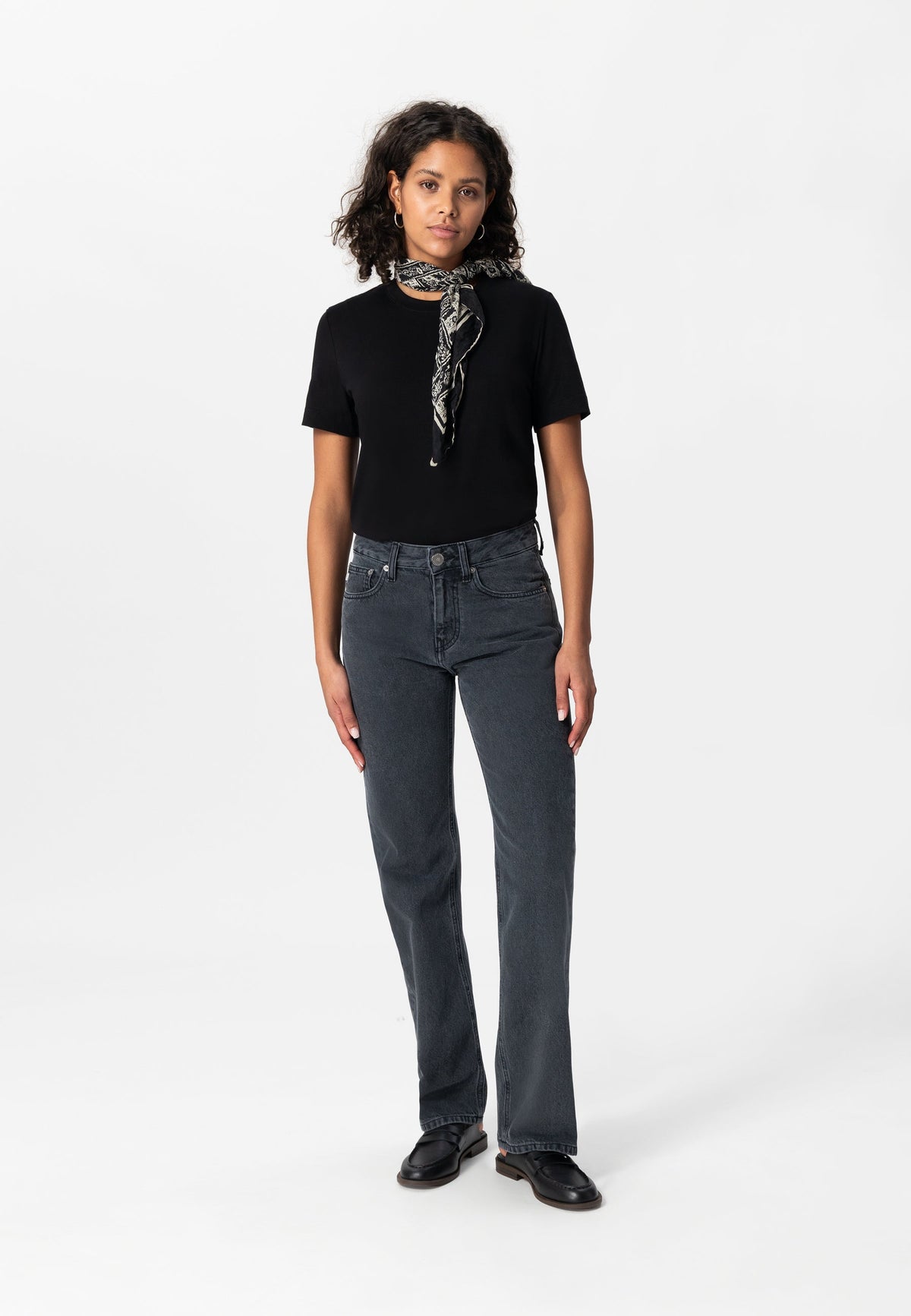 Woman wearing a black T-shirt and dark wash straight-leg jeans, styled with black loafers and a patterned neck scarf.