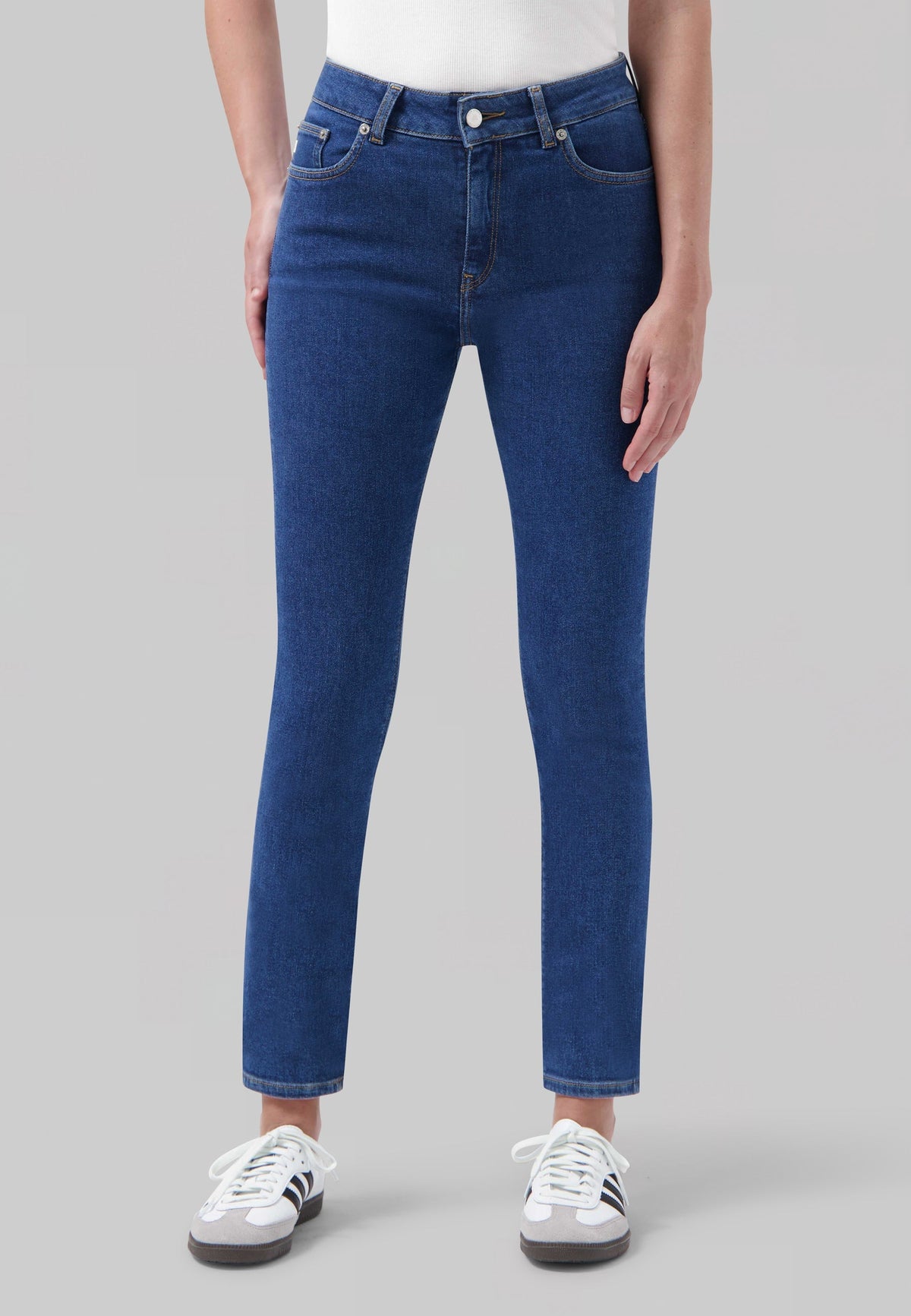 Model wearing classic blue denim jeans with high waist and slim fit, paired with casual white sneakers and a simple top.