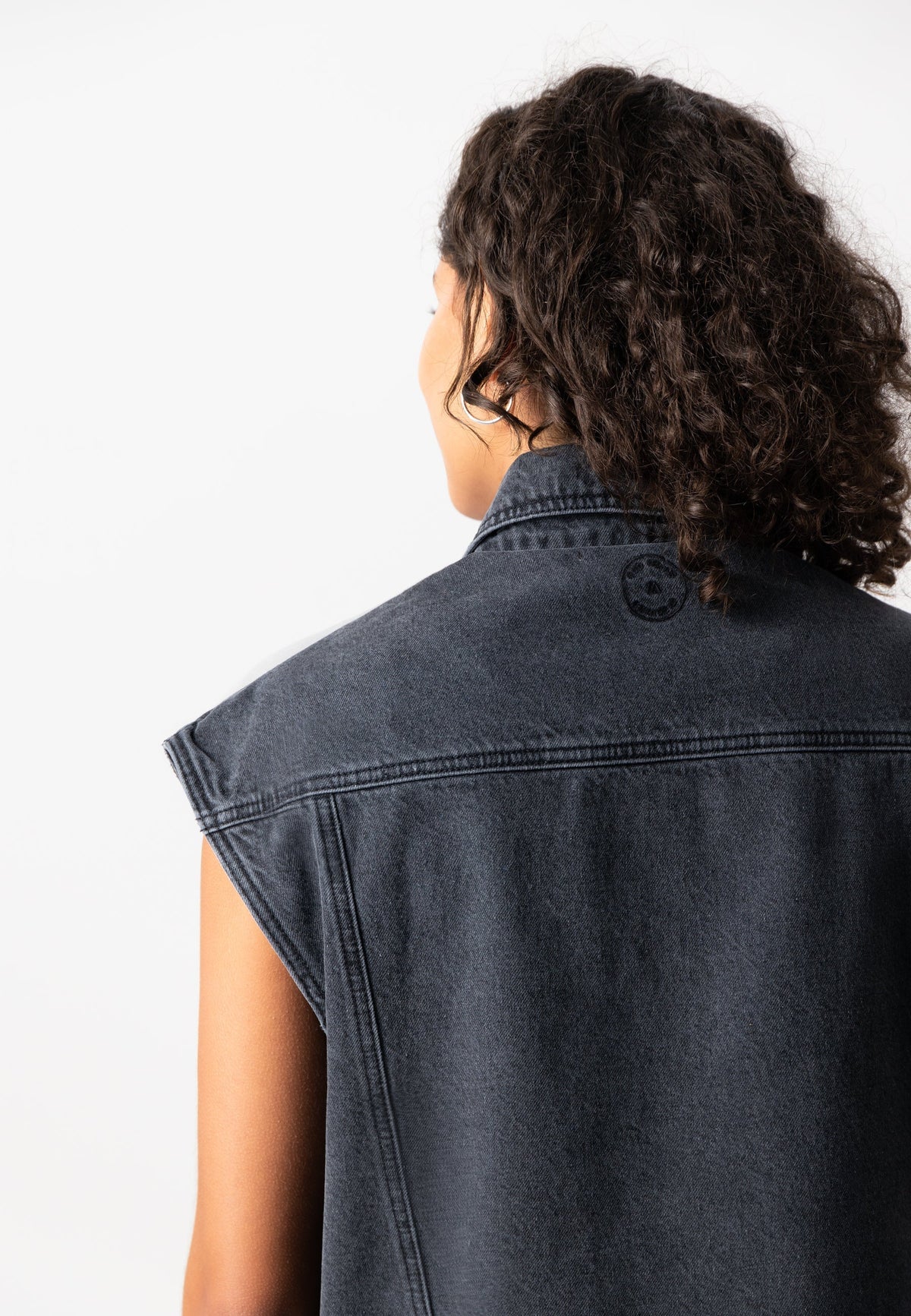 Denim sleeveless jacket in dark wash, showcasing back view with detailed stitching and collar, worn by a person with curly hair.