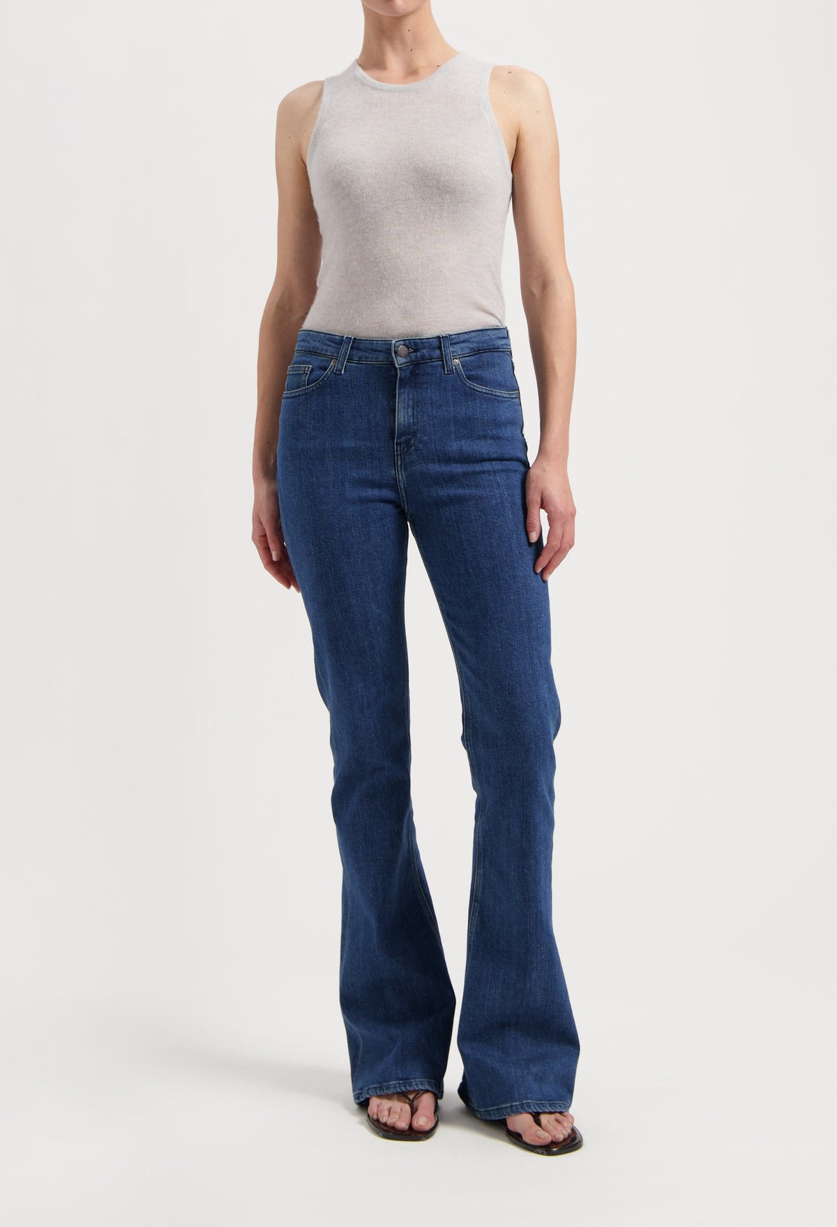 Woman wearing a light gray tank top paired with blue bootcut jeans and flip-flops, showcasing casual fashion.