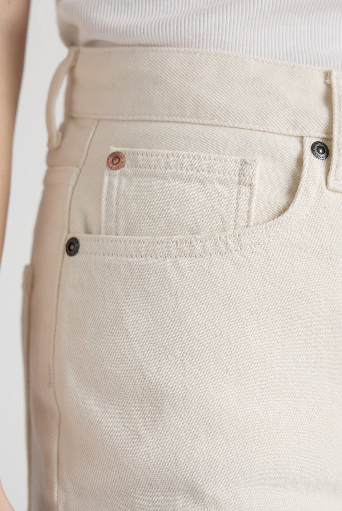 Close-up of cream-colored denim jeans pocket on a textured fabric, showing intricate stitching details and metal rivet buttons.