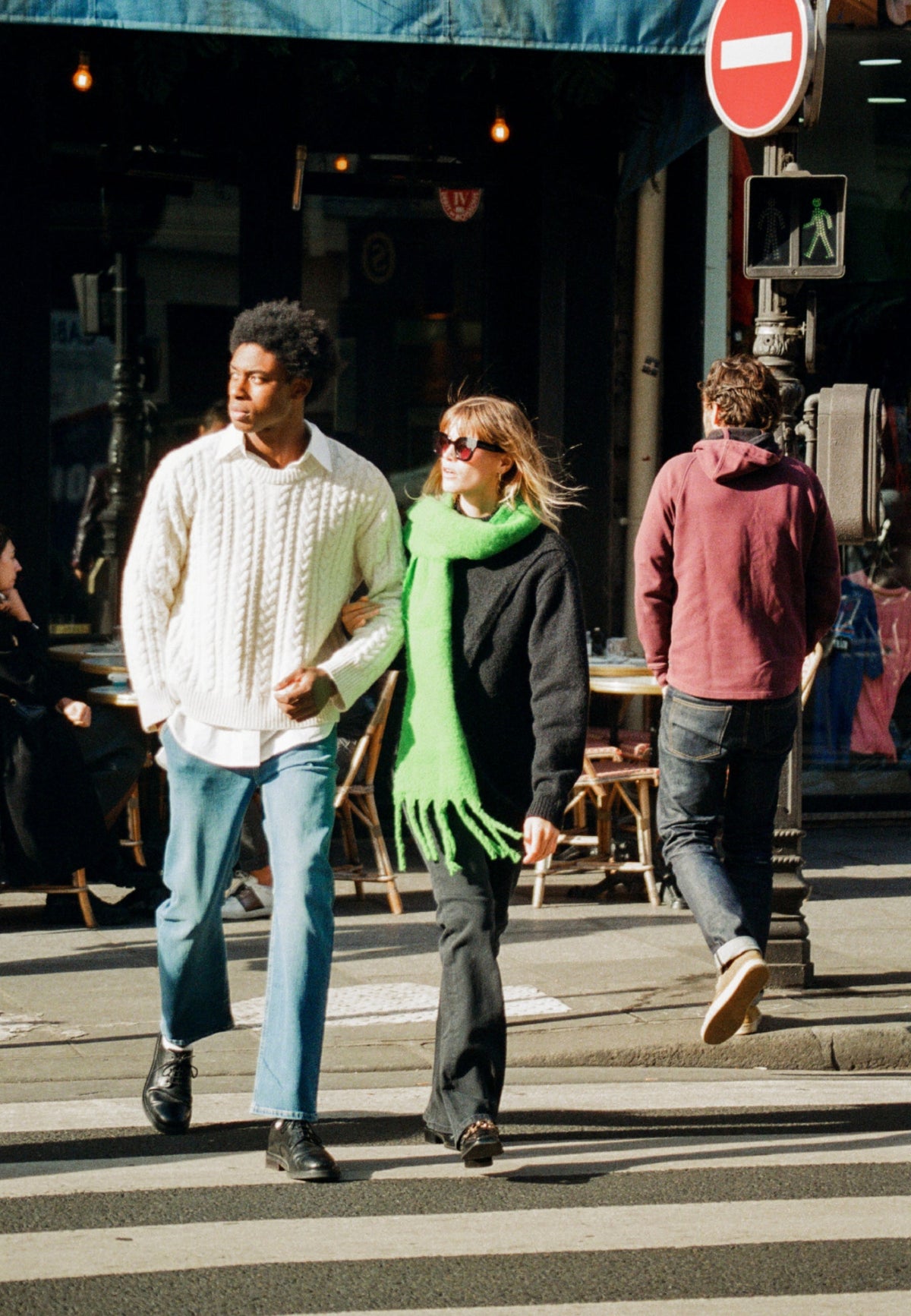Couple crossing street wearing stylish winter outfits, including a white cable knit sweater and a bright green scarf.