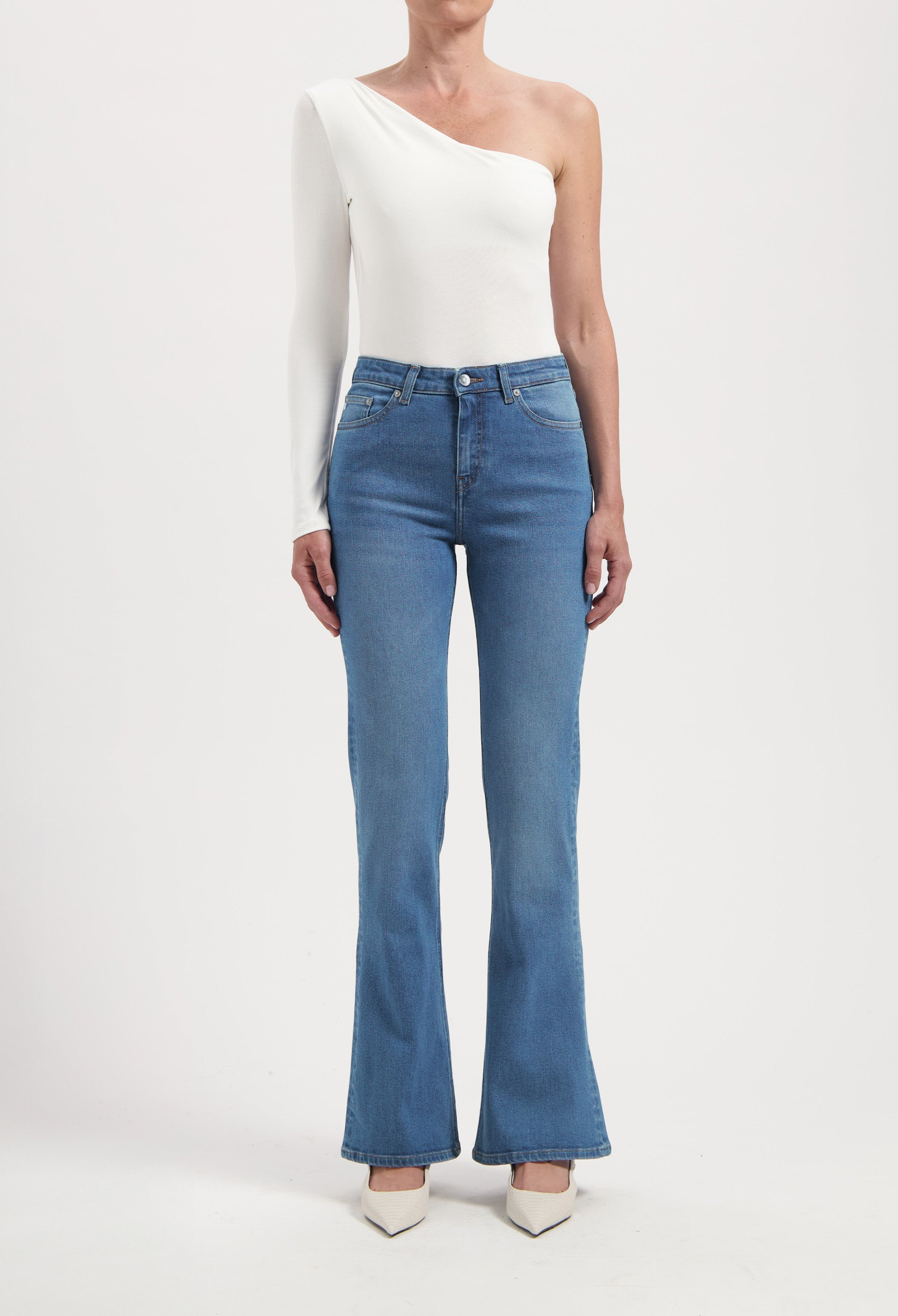 One-shoulder white top paired with blue flared jeans and white heels, showcasing minimalist fashion style.