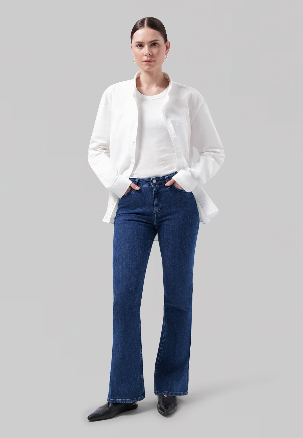 Model wearing flared dark blue jeans with a fitted white T-shirt and a casual white button-up shirt, against a neutral background.