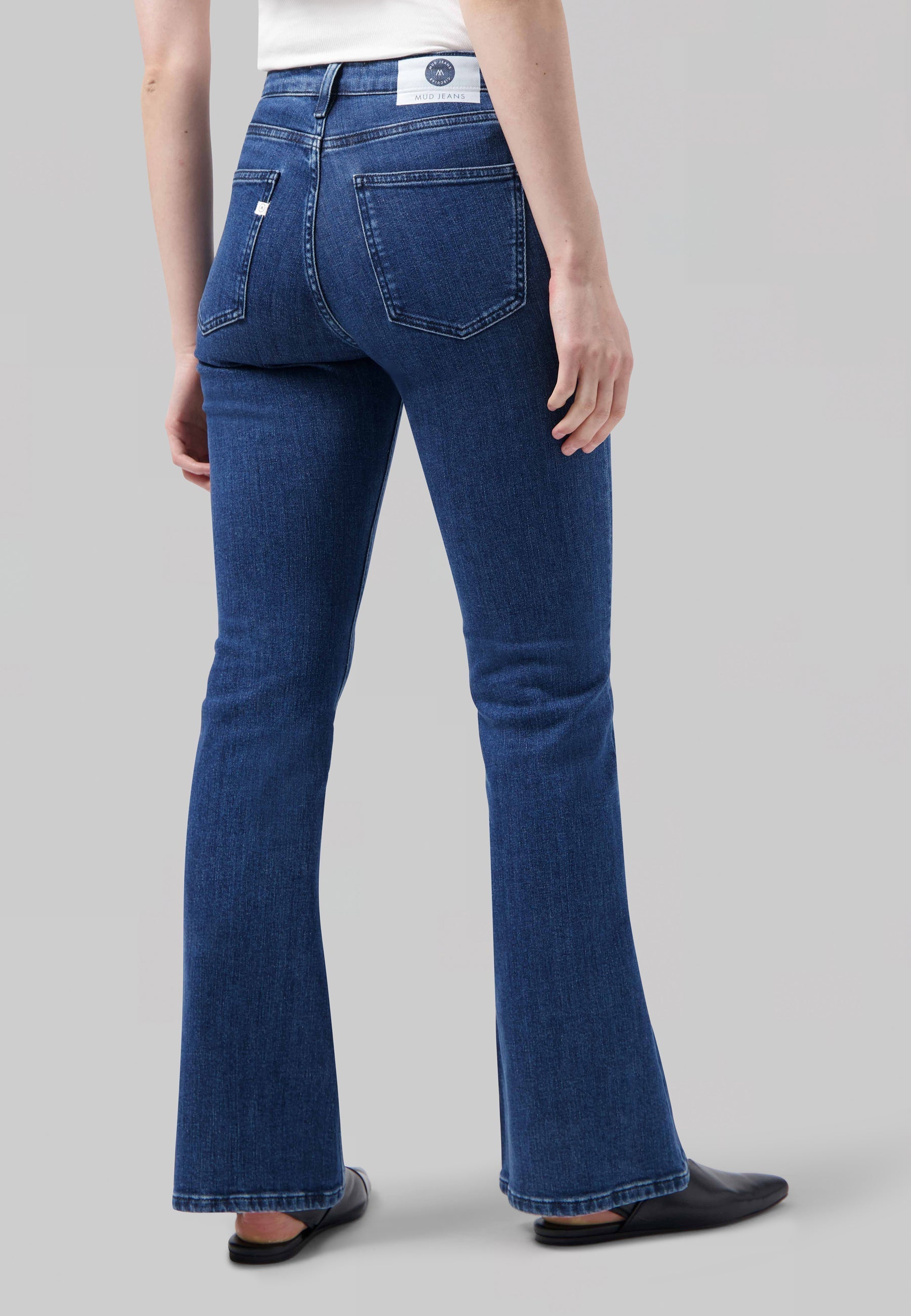 Person wearing classic bootcut blue jeans from MUD Jeans, rear view focusing on denim texture and fit, paired with black shoes.