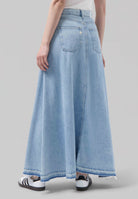 Light blue denim maxi skirt with frayed hem, high-waisted design, and back pockets, styled with white sneakers.
