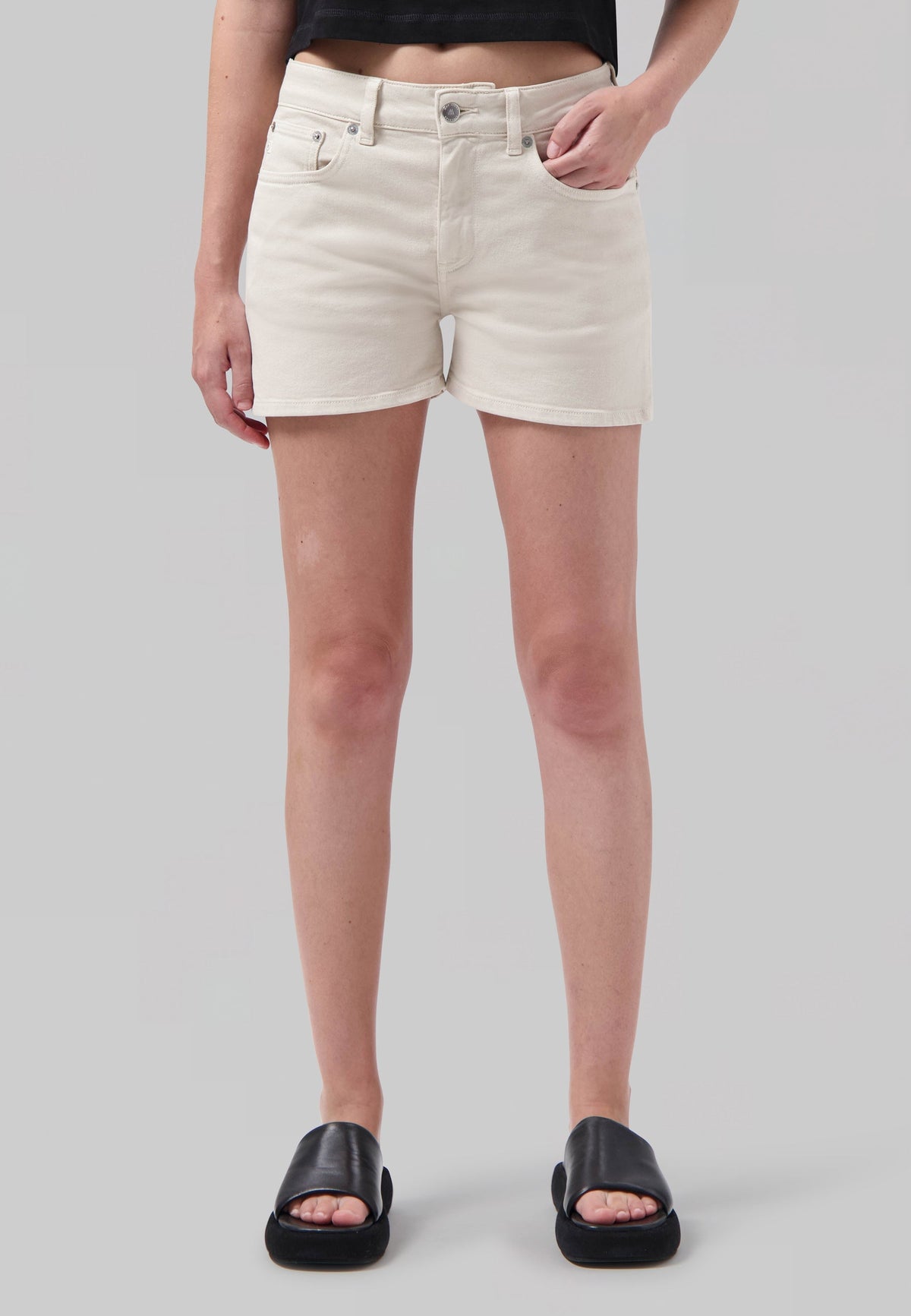 Woman wearing off-white denim shorts with a high-waist fit, styled with a cropped black top and black leather slide sandals.