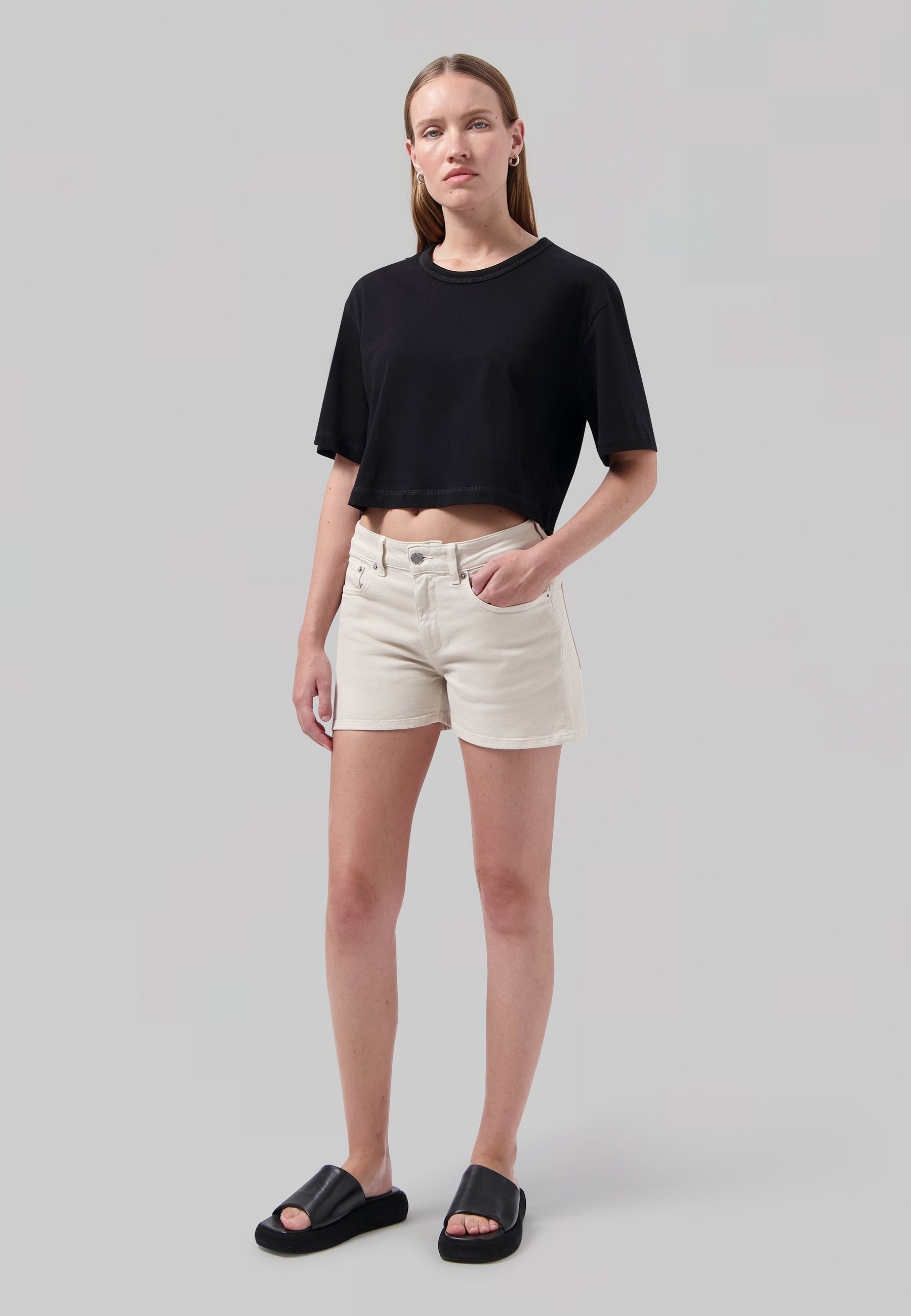 Casual outfit featuring a woman in a black short-sleeve crop top and beige shorts, paired with black slide sandals on a plain background.