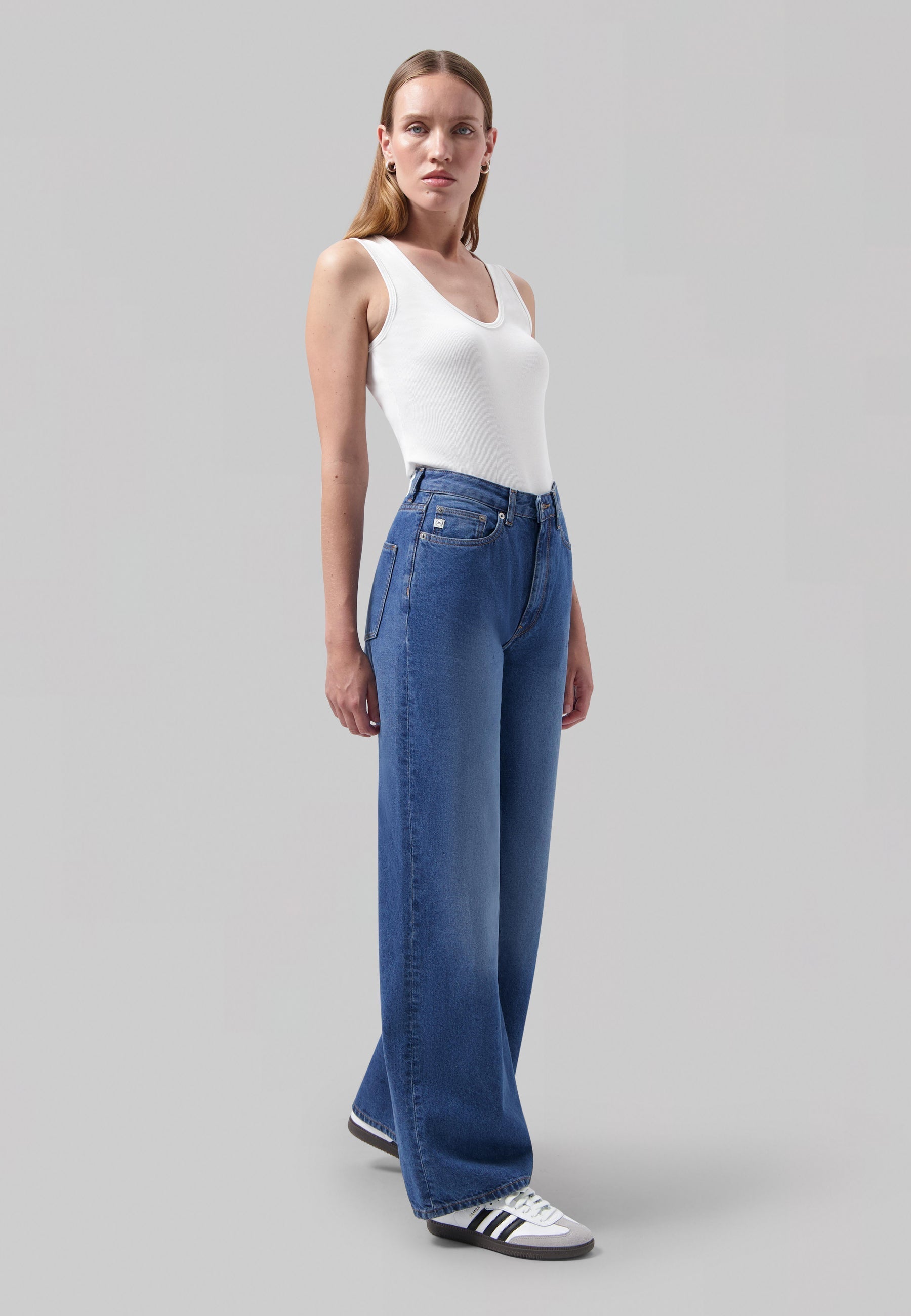White sleeveless top paired with wide-leg blue jeans and classic black-and-white sneakers on a neutral backdrop.