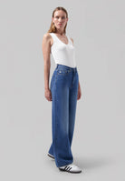 White sleeveless top paired with wide-leg blue jeans and classic black-and-white sneakers on a neutral backdrop.