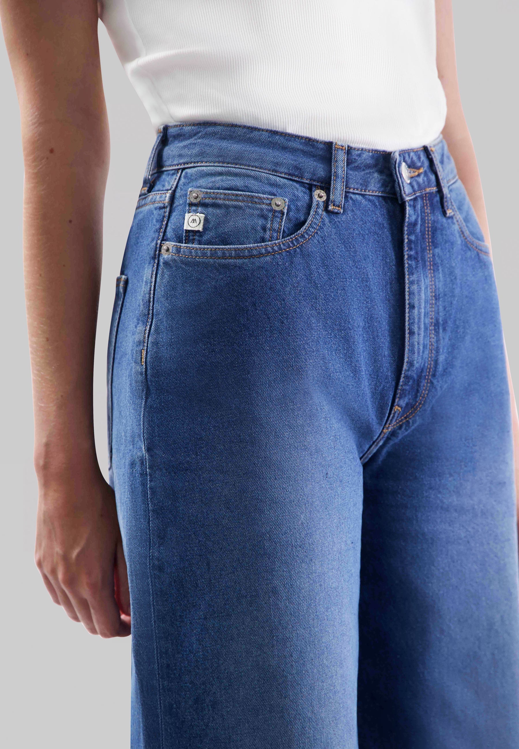 High-waisted blue jeans with a fitted waist and button fly, worn with a sleeveless white ribbed top.