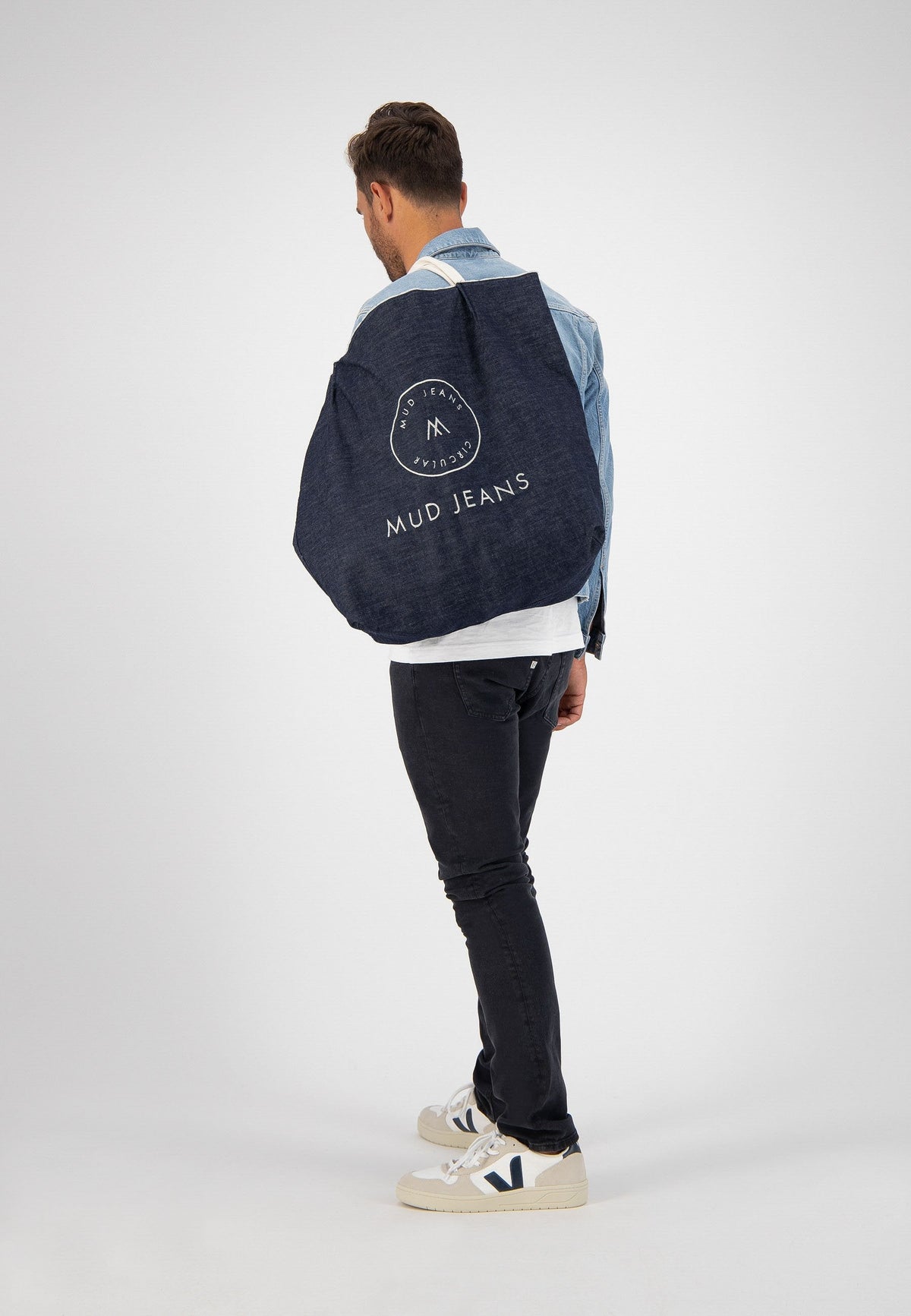 Man wearing a light blue denim jacket and dark jeans with a large blue MUD Jeans tote bag slung over his shoulder.