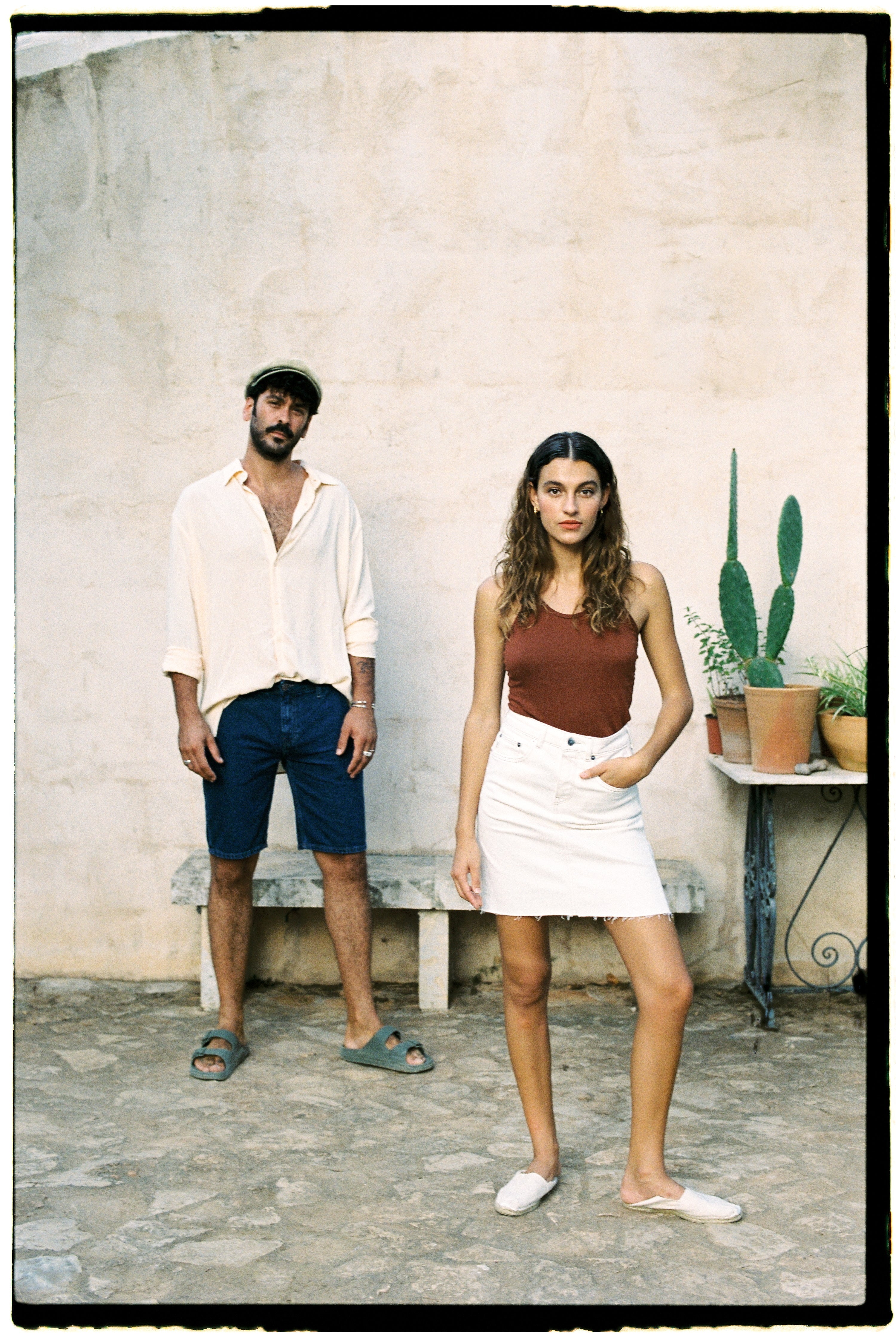 Casual summer fashion with a beige shirt, navy shorts, and sandals for him, and a brown top with a white skirt for her.