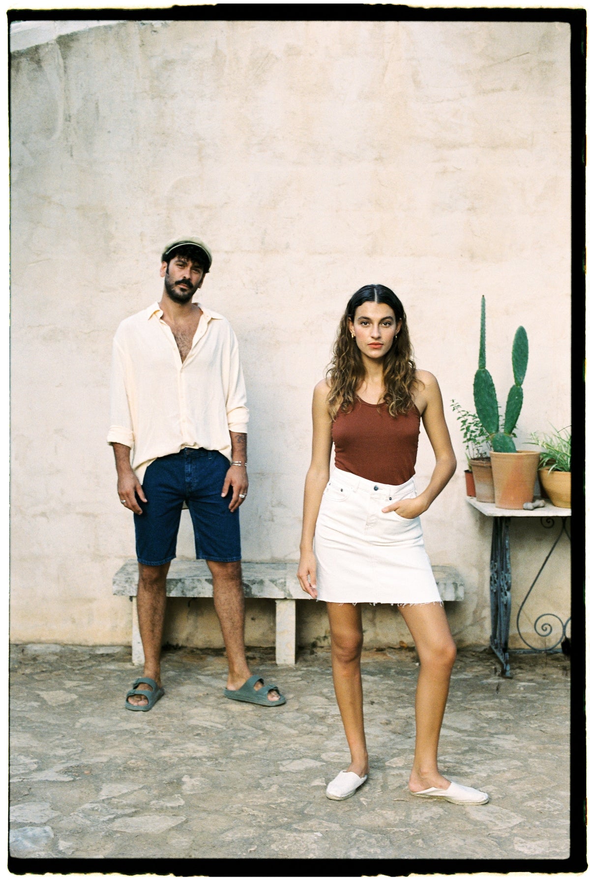 Casual summer fashion with a beige shirt, navy shorts, and sandals for him, and a brown top with a white skirt for her.