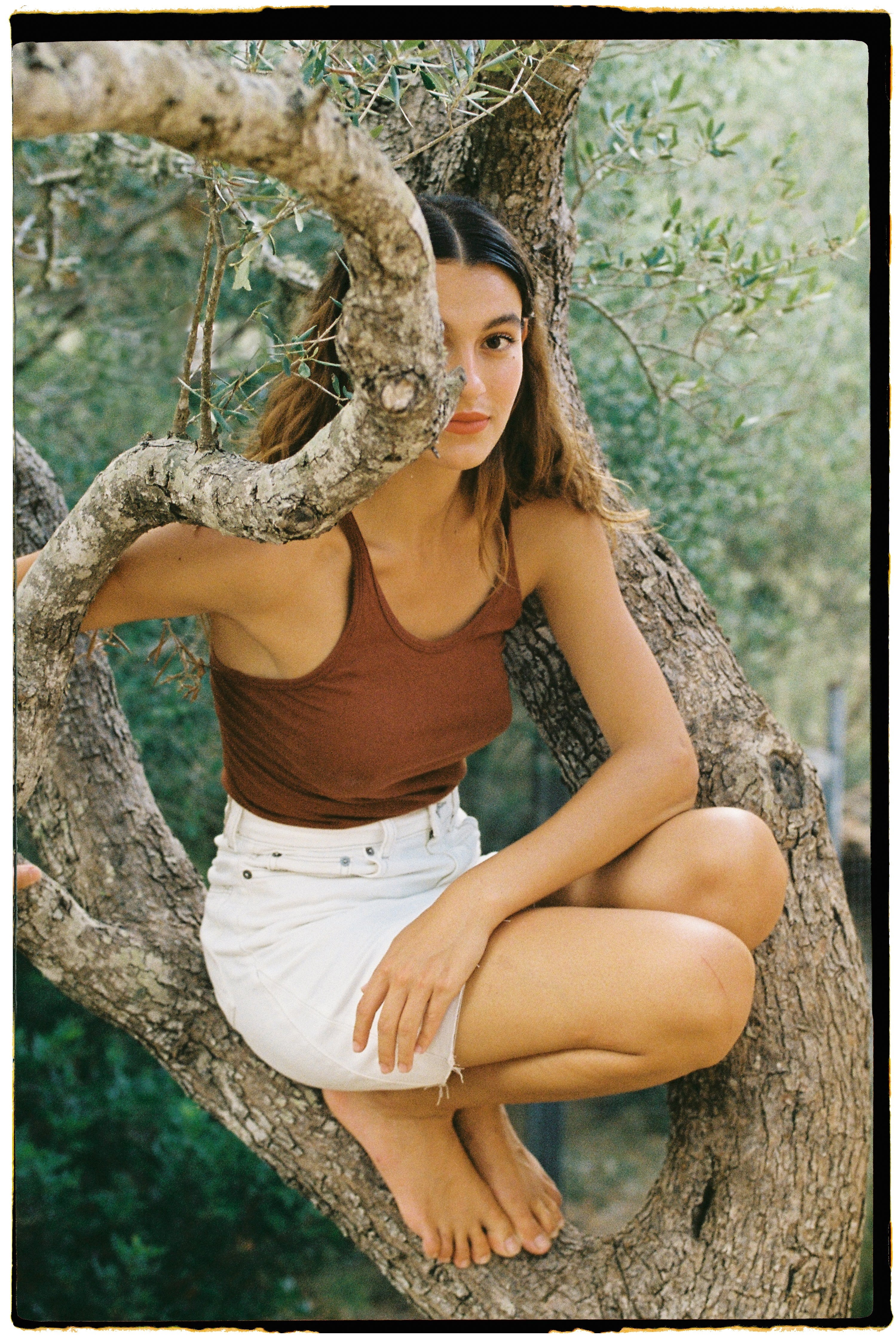 Woman in a brown tank top and white denim skirt sitting on a branch of an olive tree in a natural setting.