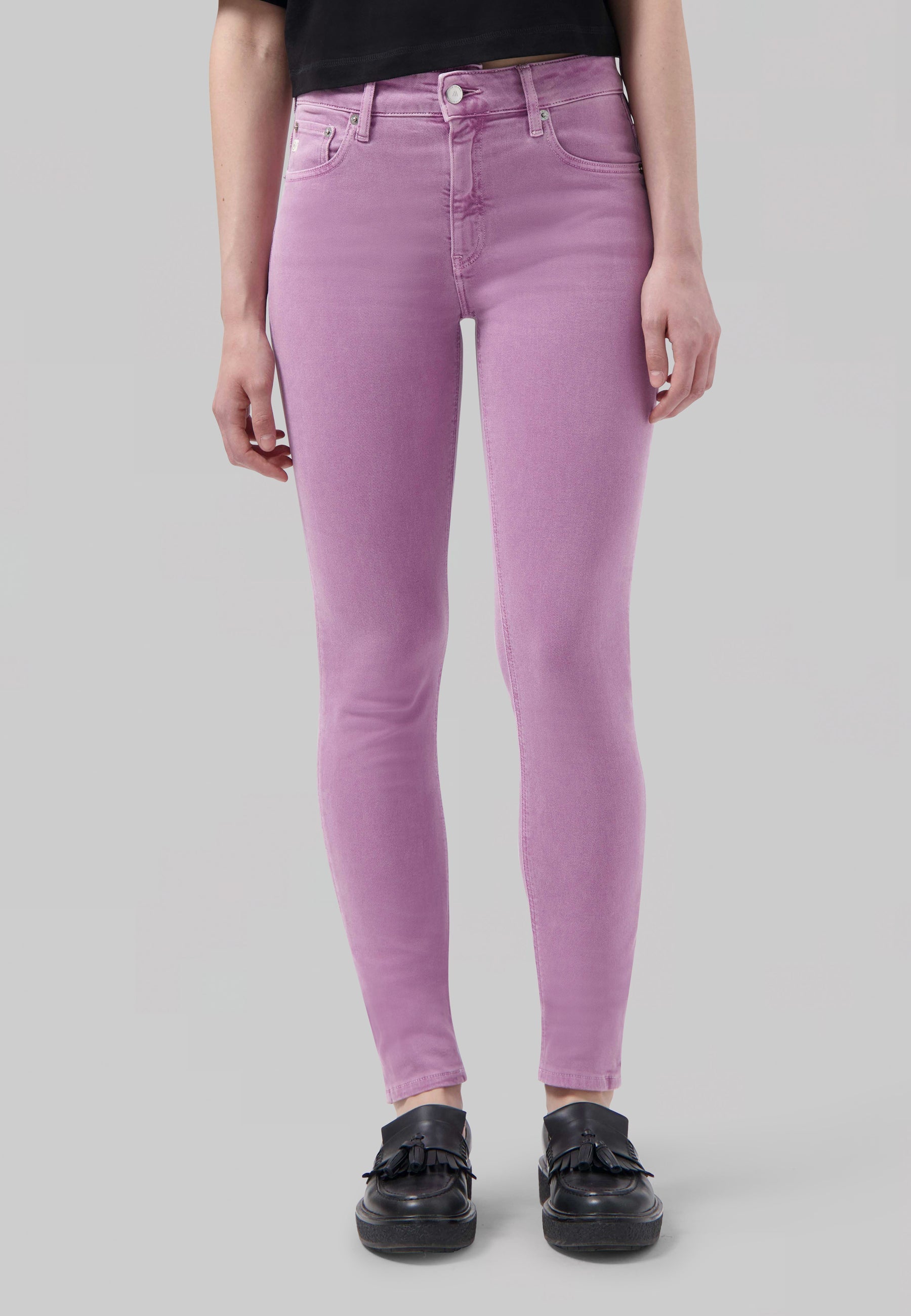 Woman wearing pastel purple, high-waisted skinny jeans paired with a black top and black loafers in a studio setting.