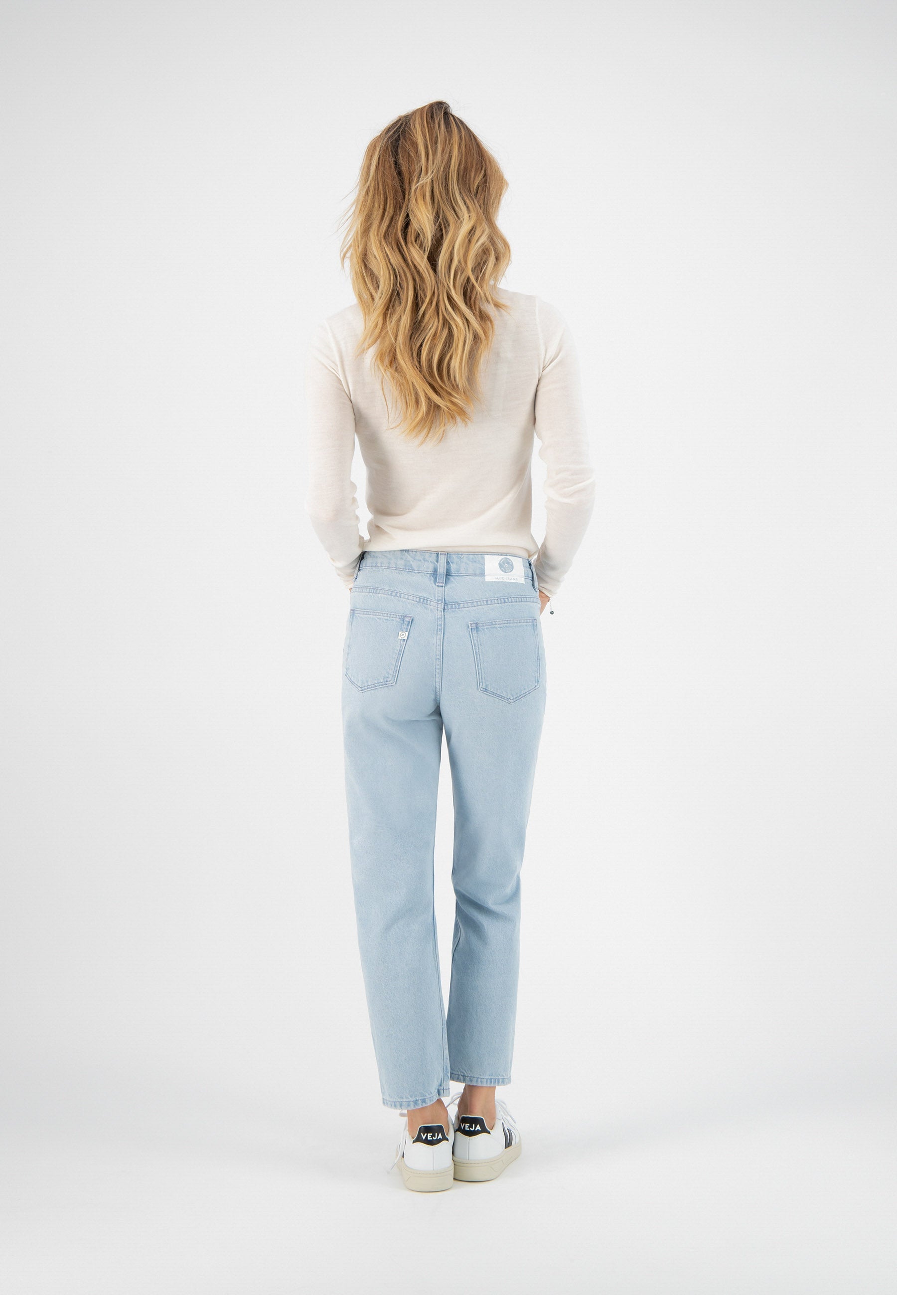 Blonde woman in a white long-sleeve top and light blue jeans, standing with back view, wearing white sneakers.