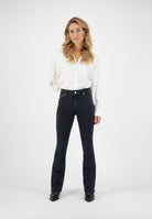 A woman wearing a white button-up blouse and black flared jeans paired with black pointed-toe boots against a plain background.