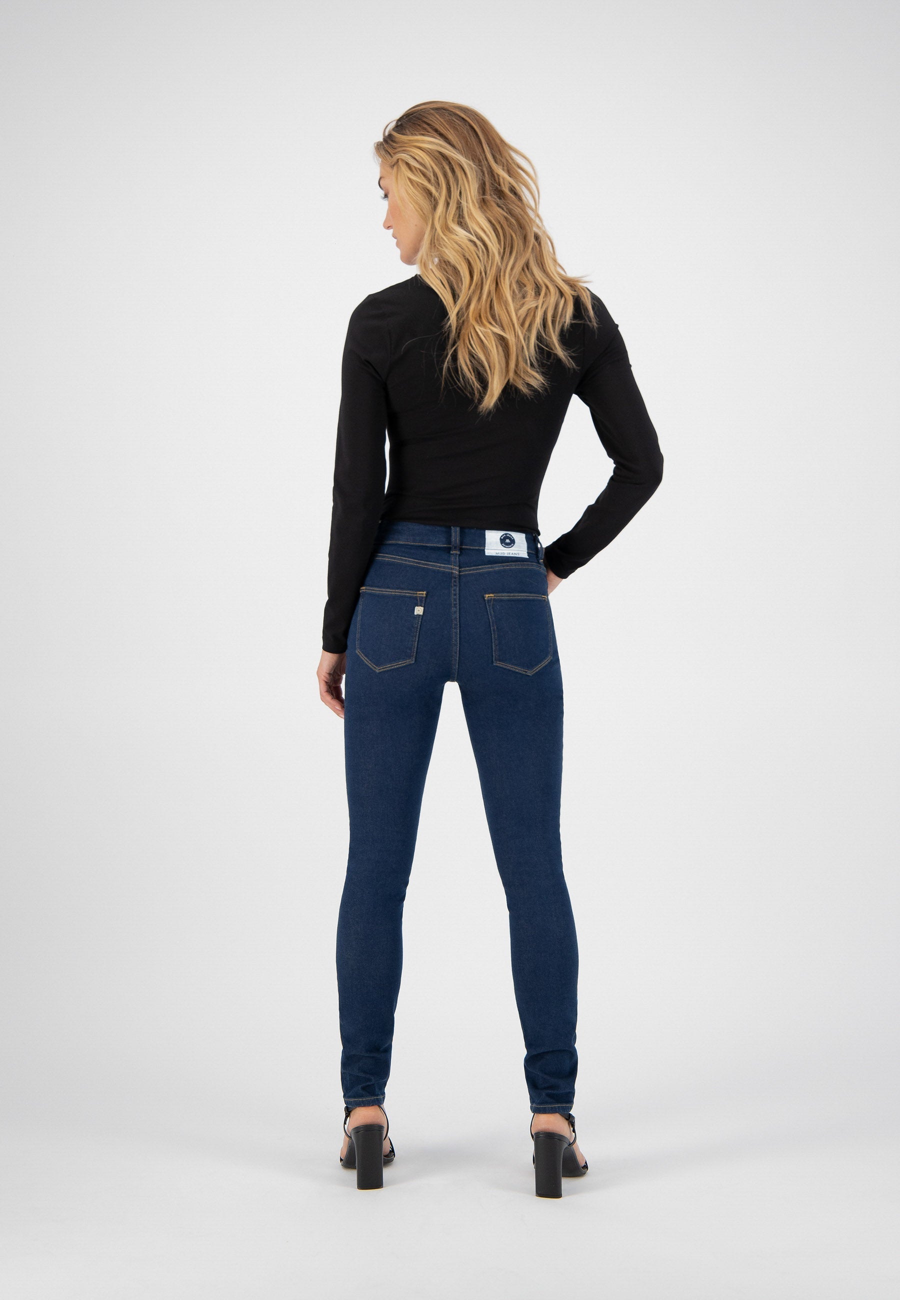Blonde woman poses in dark blue, slim-fit jeans paired with a long-sleeve black top, showcasing the back design.