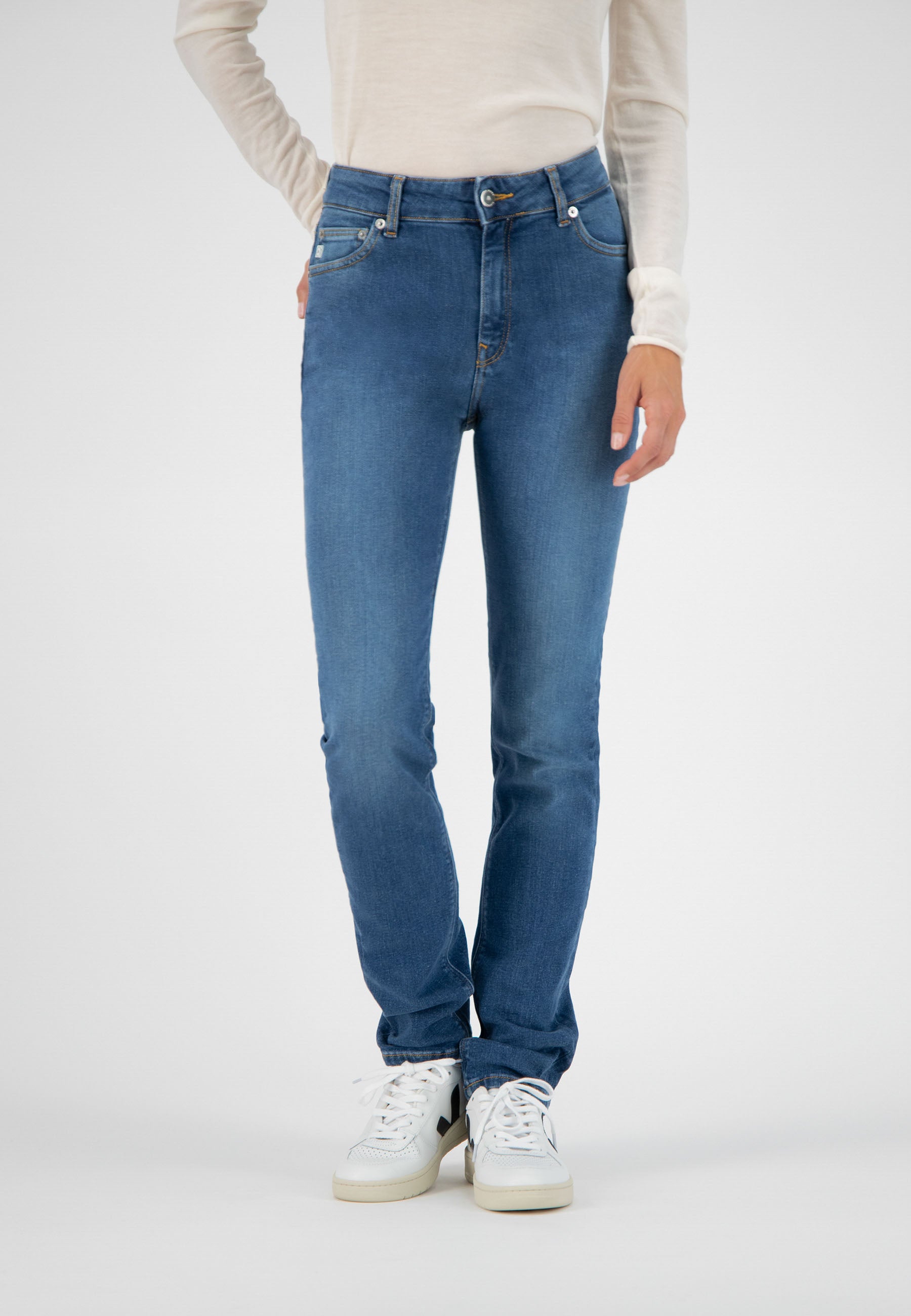 Woman wearing blue straight-fit jeans with white sneakers and a light beige long-sleeve top, highlighting casual style.