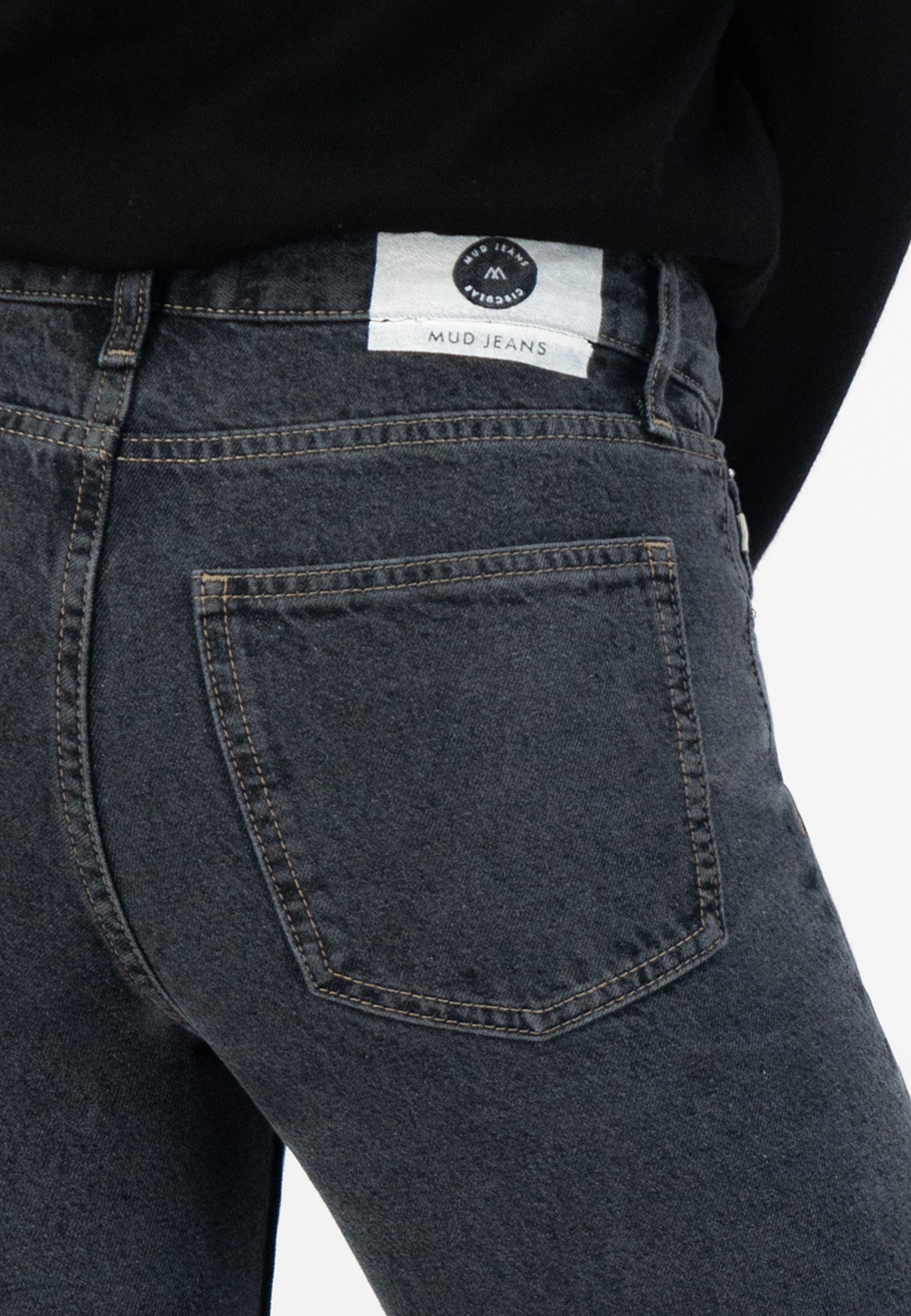 Dark gray MUD Jeans in organic cotton, highlighting close-up of back pocket stitch detail, worn with a black top.
