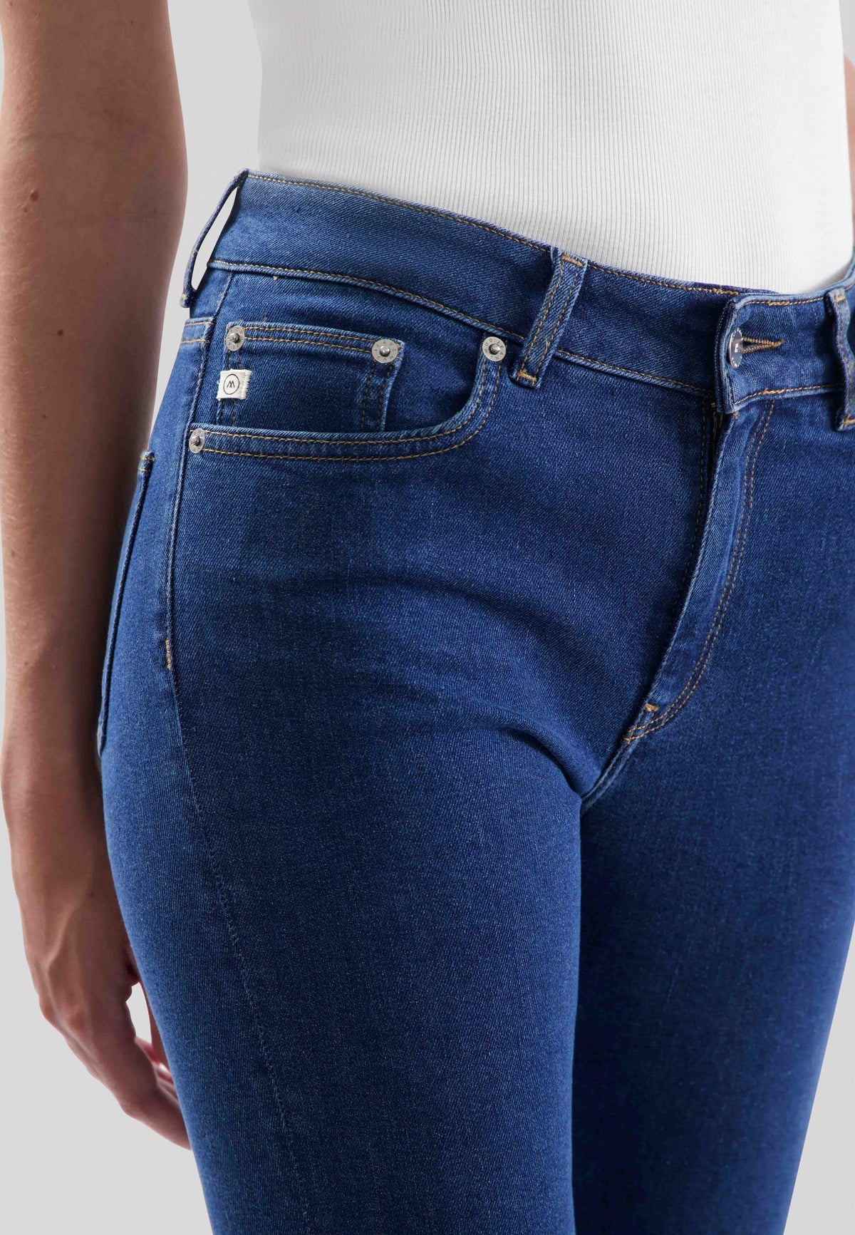 Blue slim-fit denim jeans in organic cotton, close-up view on waist and pockets, paired with a white ribbed top.