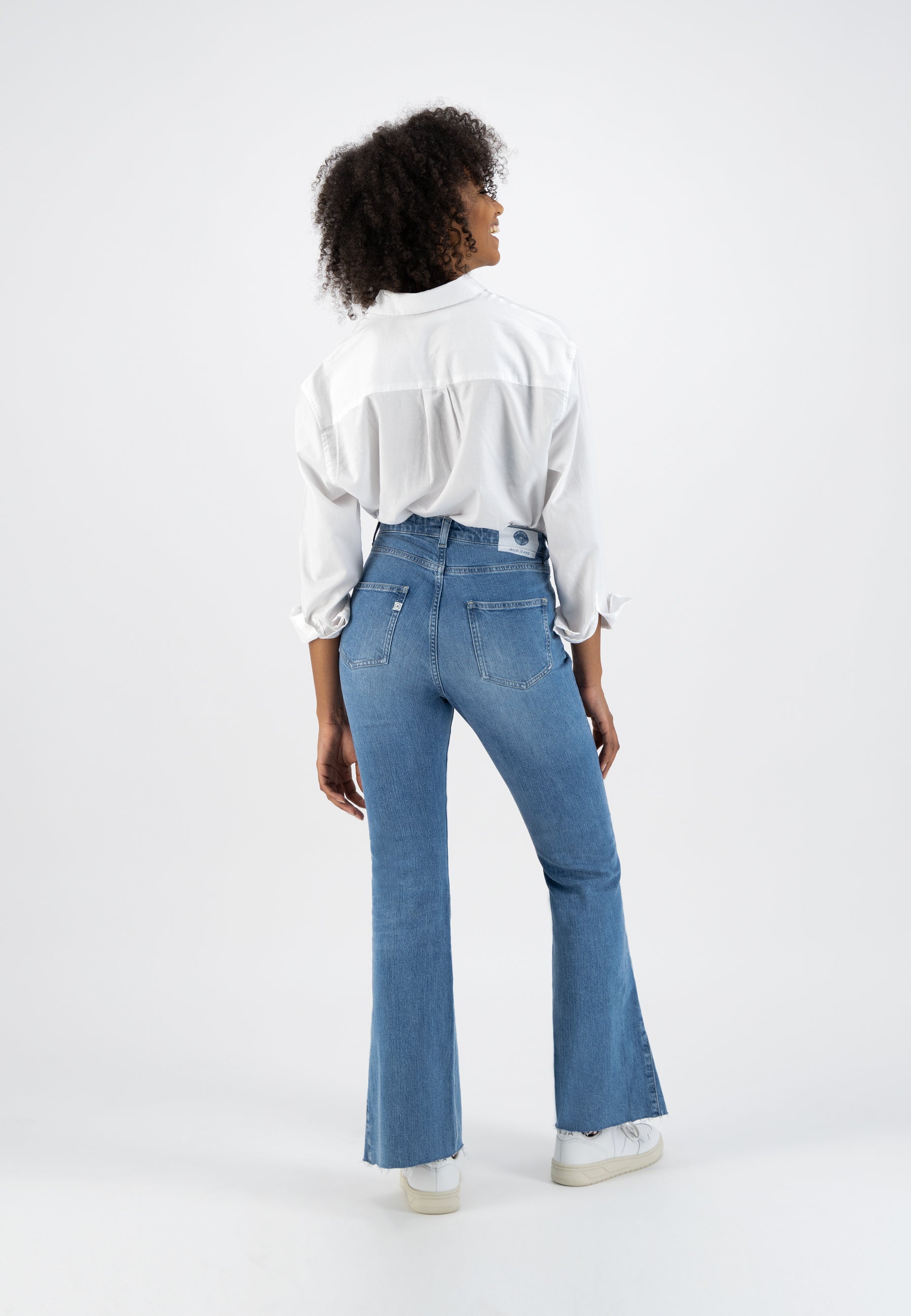 Model wearing a white button-up shirt and high-waisted blue flare jeans, with sneakers, viewed from the back.
