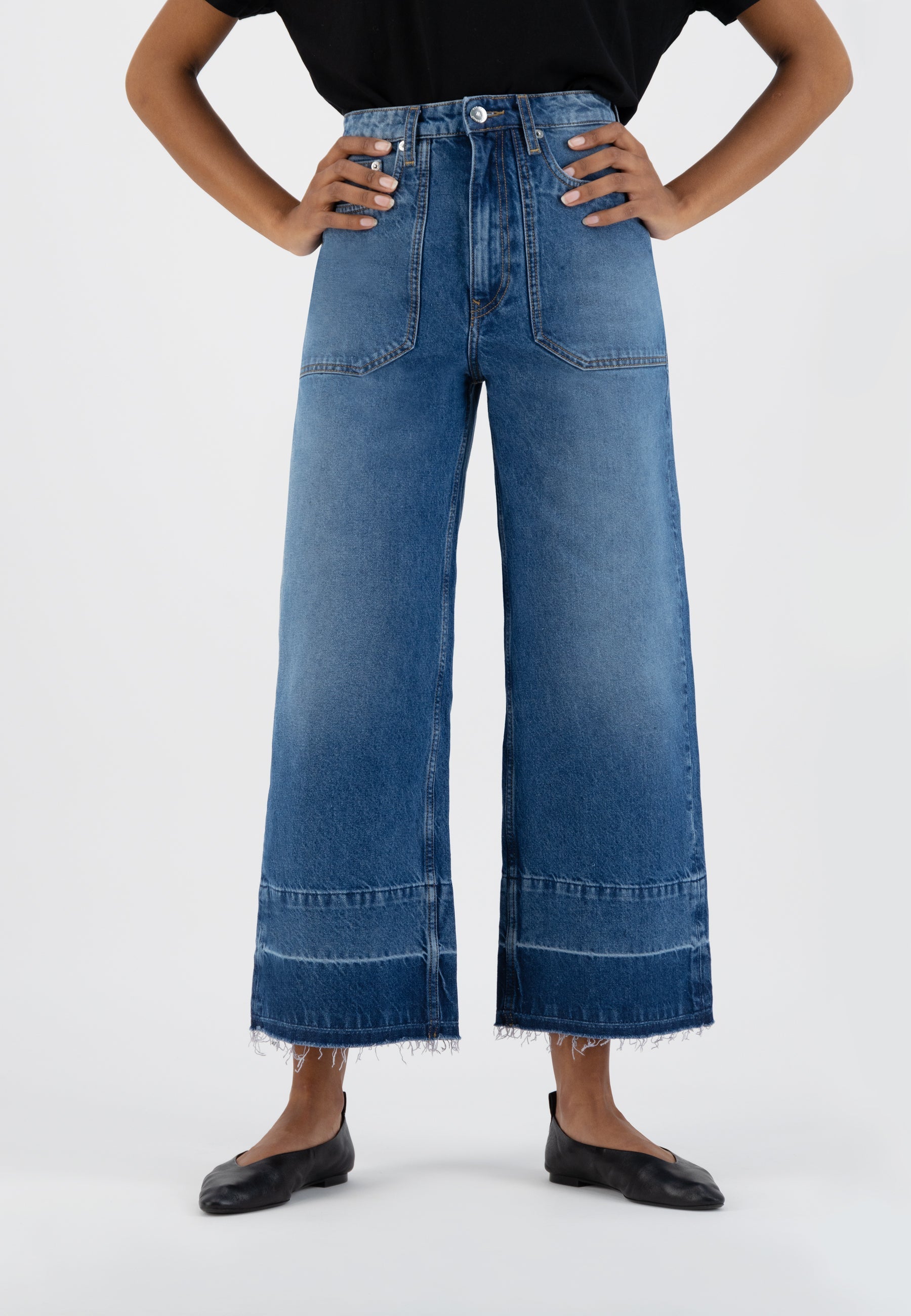 High-waisted wide-leg denim jeans in medium blue with front pockets, worn with a black top and flat black shoes.