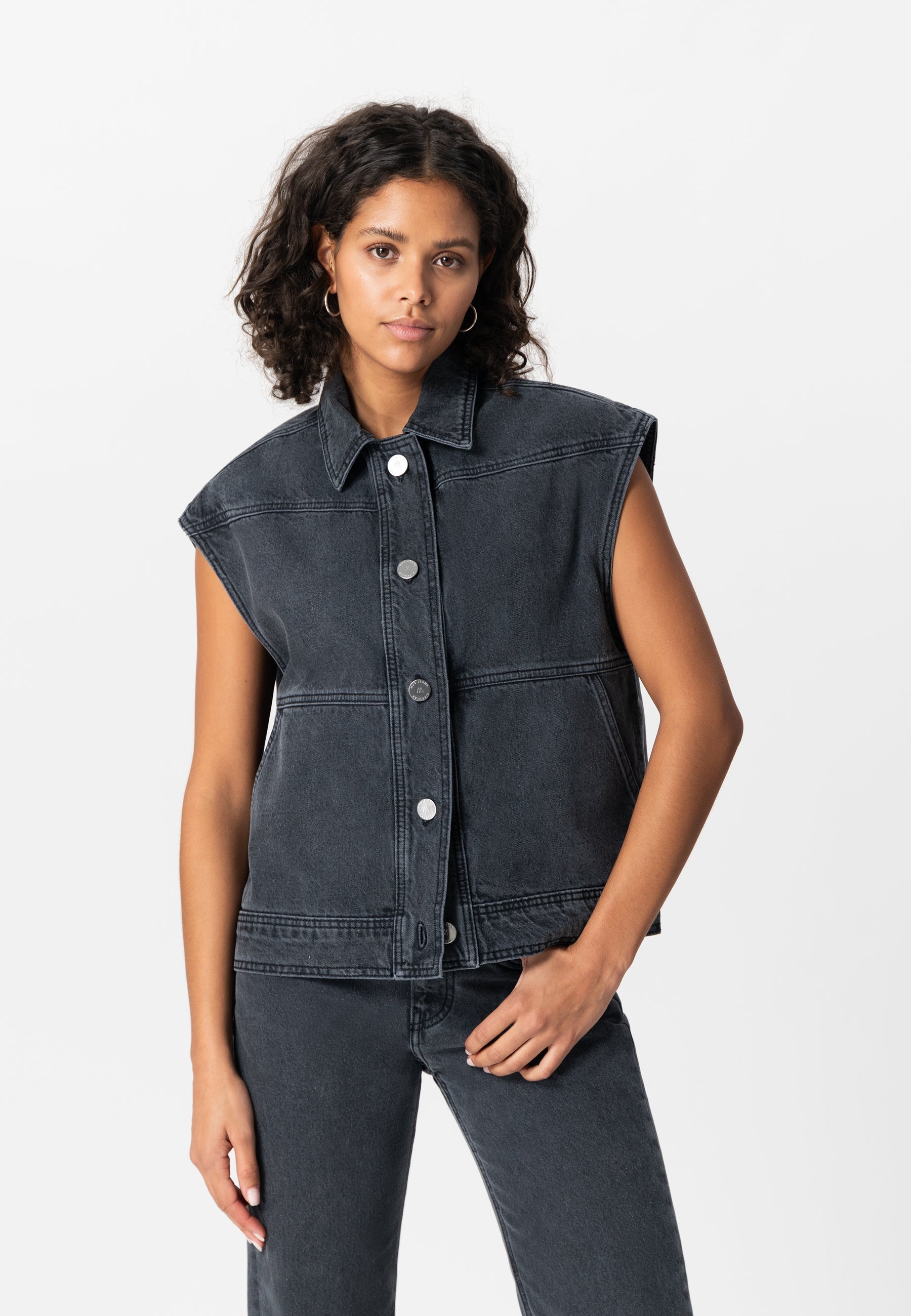 Sleeveless denim jacket in washed black with button closure, styled with matching high-waisted jeans, worn by model.