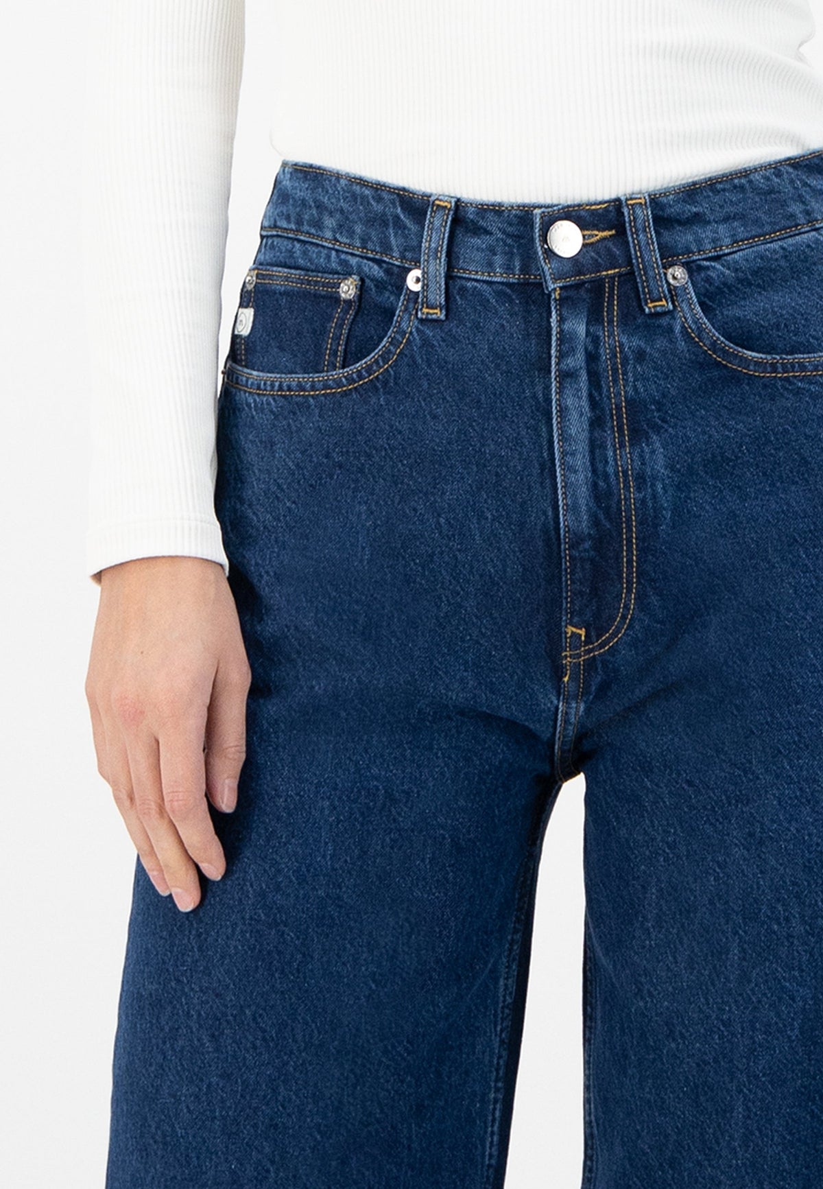 Close-up of blue high-waisted jeans with yellow stitching, worn with a white ribbed long-sleeve top.