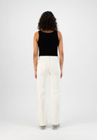 Curly-haired woman in a black sleeveless top and white high-waisted, wide-leg trousers, facing away in a minimalist setting.
