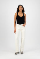Woman wearing a black sleeveless top paired with high-waisted, off-white denim jeans and gray sneakers against a white background.