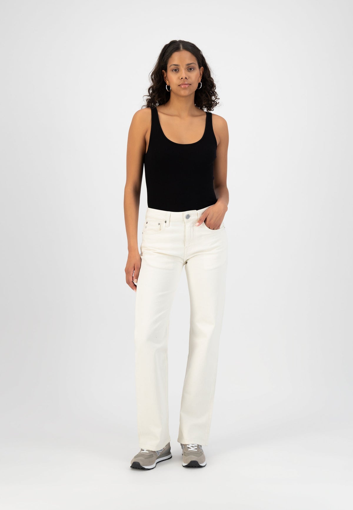 Woman wearing a black sleeveless top paired with high-waisted, off-white denim jeans and gray sneakers against a white background.