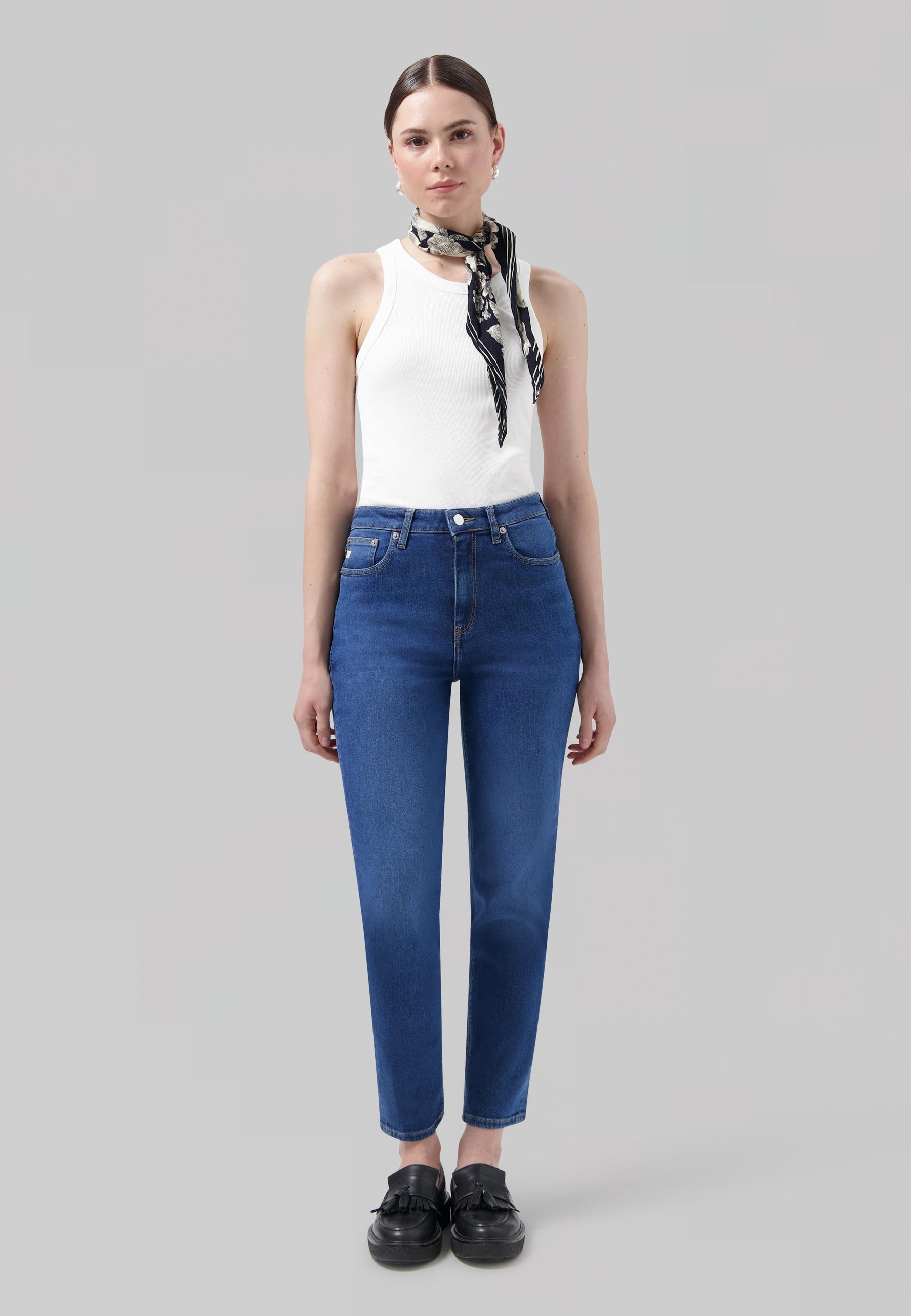 A woman wearing blue high-waisted jeans and a white sleeveless top with a black and white patterned scarf.