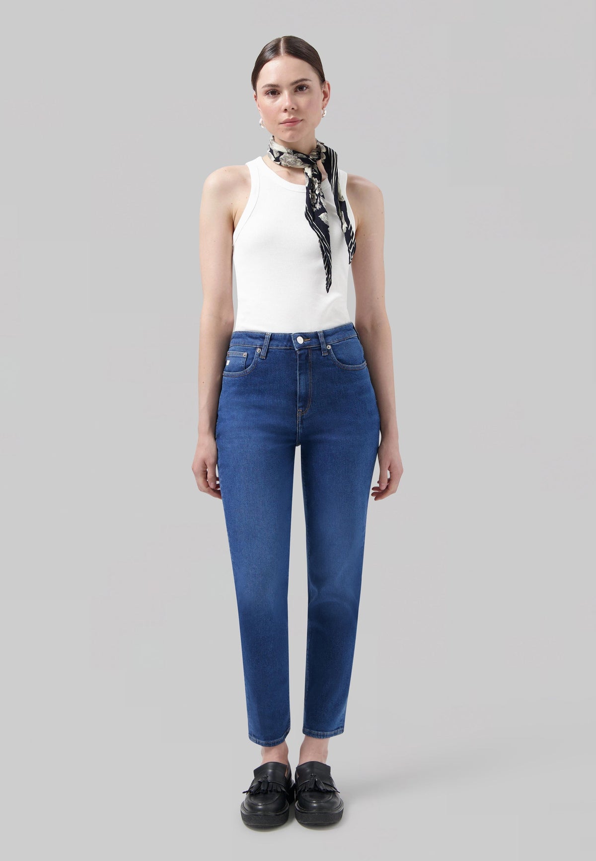 A woman wearing blue high-waisted jeans and a white sleeveless top with a black and white patterned scarf.