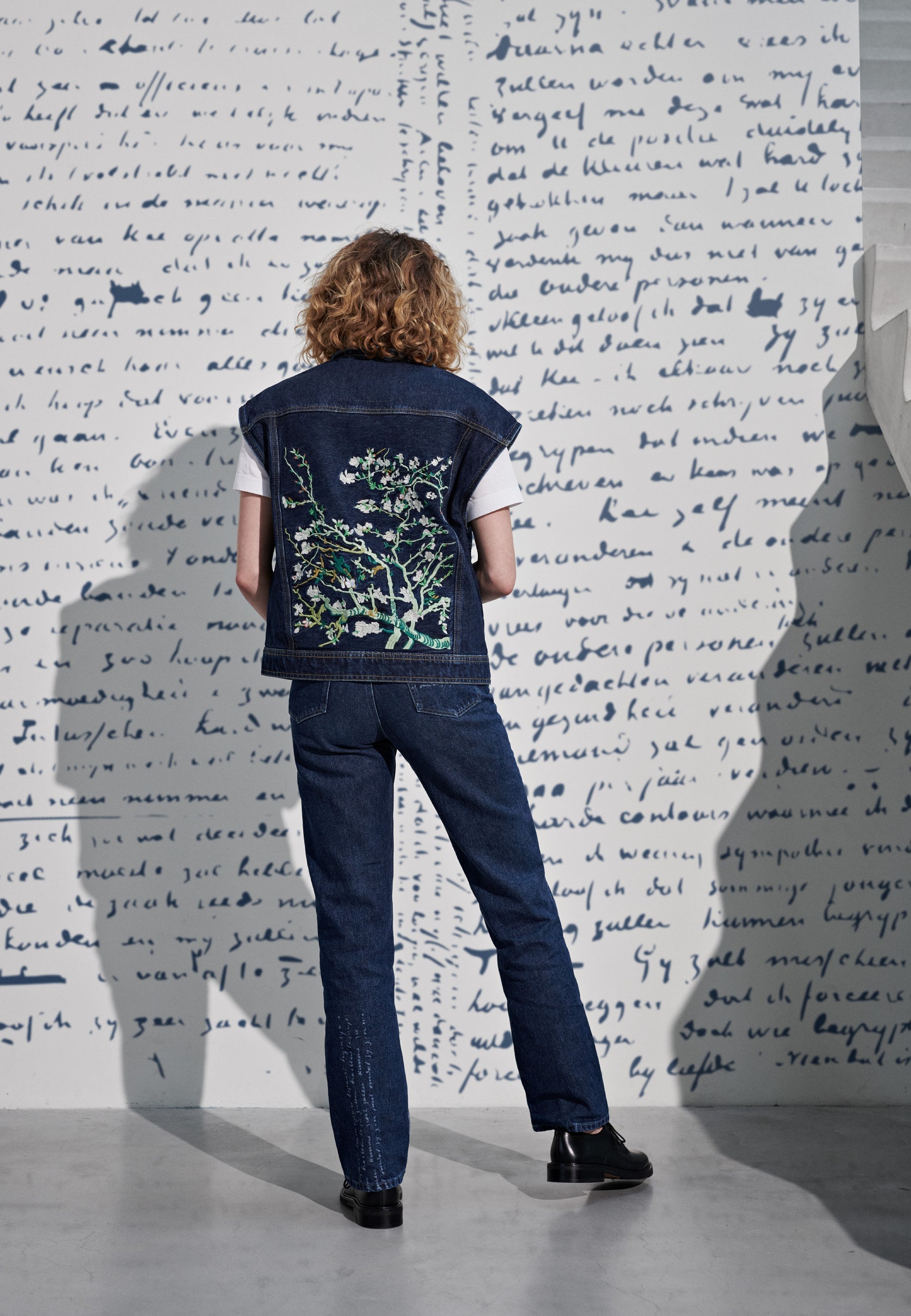 Curly-haired person wearing embroidered dark denim vest and jeans, standing against a wall with handwritten script design.