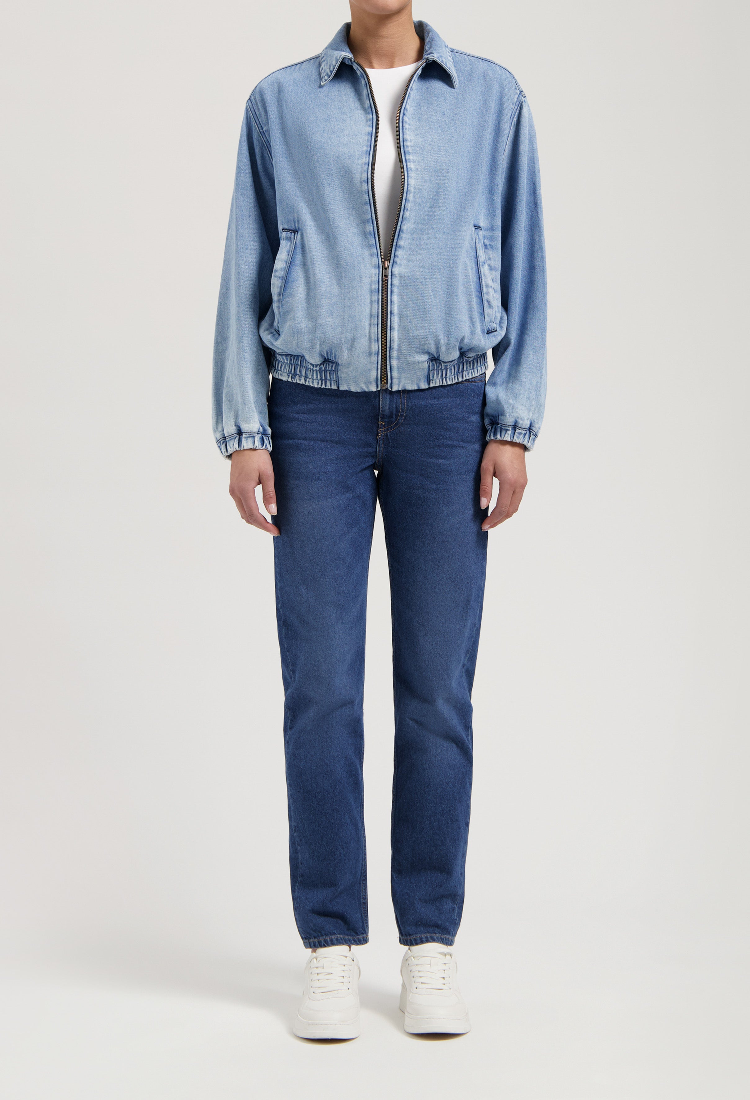 Person wearing a light blue denim jacket with front zipper, paired with dark blue straight-leg jeans and white sneakers.