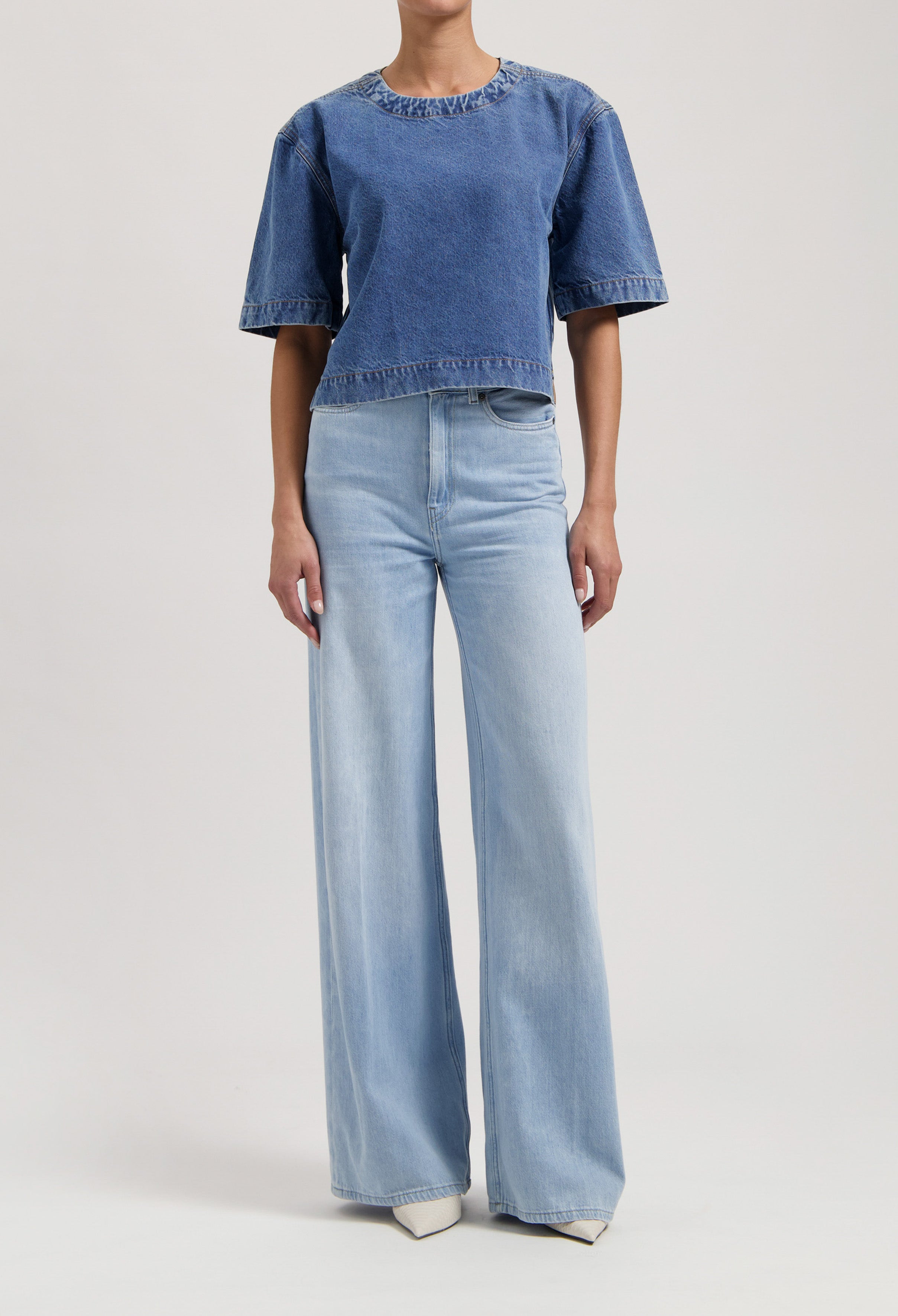 Model in wide-leg, light blue high-waisted jeans and short-sleeve dark blue denim top against plain background.