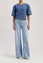 Model in wide-leg, light blue high-waisted jeans and short-sleeve dark blue denim top against plain background.